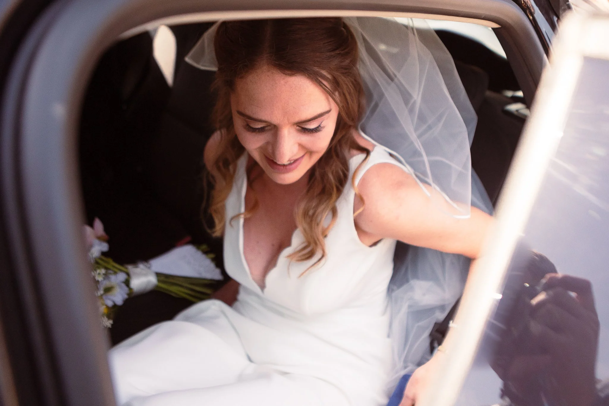 A smiling woman in a wedding dress sitting in the back of a car, holding a bouquet of flowers.