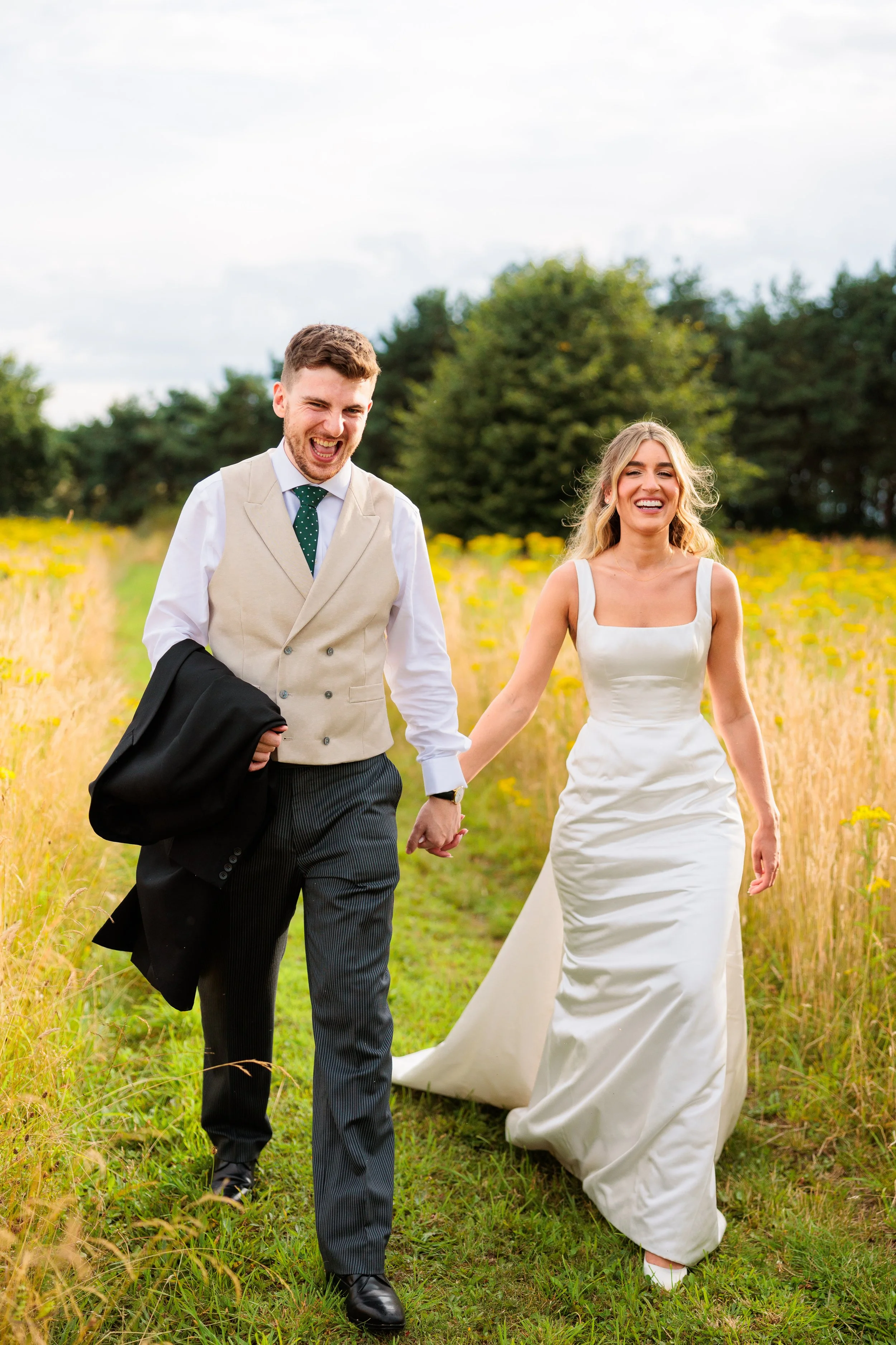 A newlywed couple walking hand-in-hand through a field with tall grass and yellow wildflowers, smiling and enjoying a sunny day outdoors.
