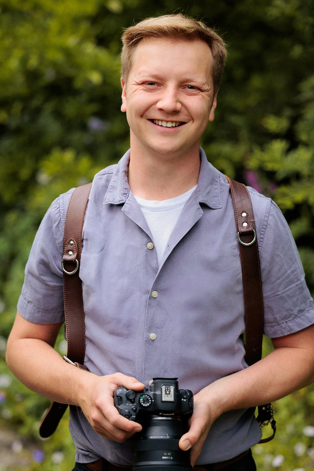 A smiling young man with short light brown hair, wearing a light gray short-sleeved shirt and carrying a brown backpack, holding a Canon camera with a large lens, standing outdoors with green foliage in the background.