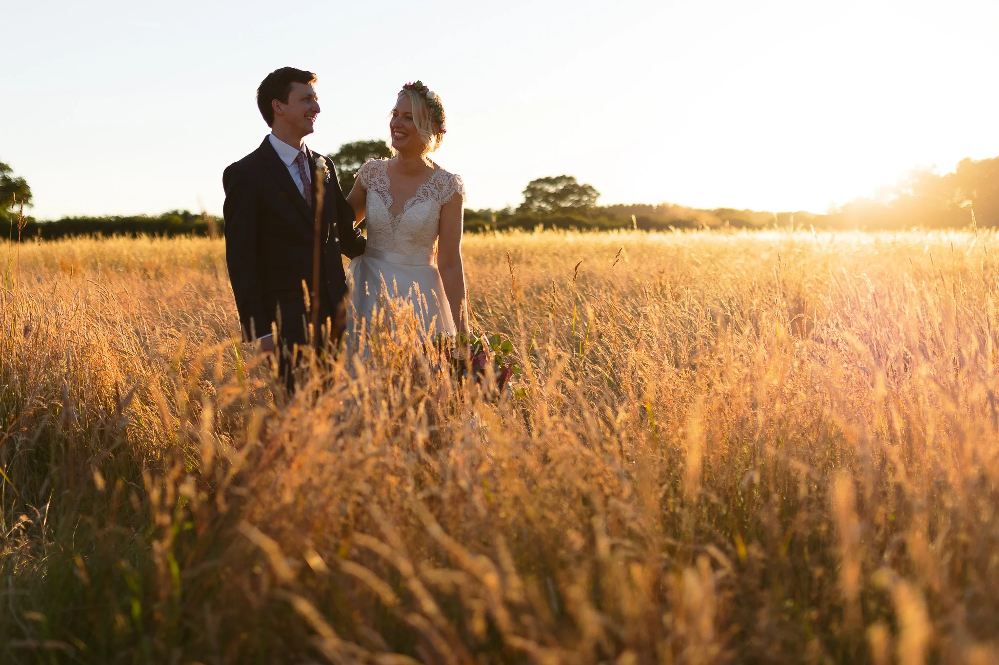 A newlywed couple stands in a sunlit golden field at sunset, smiling at each other. The bride wears a white lace wedding dress with a floral crown, and the groom is dressed in a dark suit with a tie.