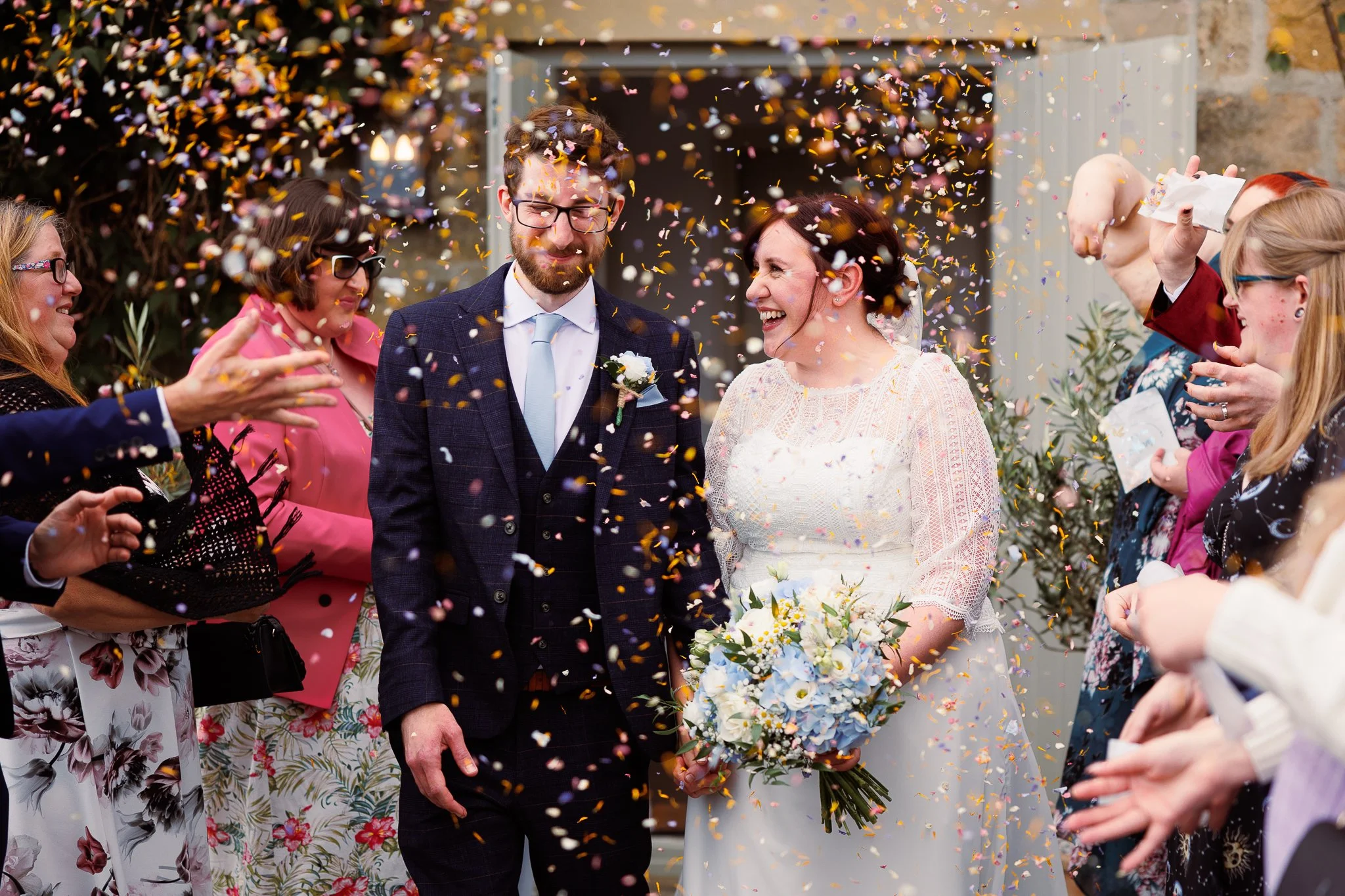 A bride and groom walking through a shower of colorful confetti surrounded by guests celebrating outside a building.