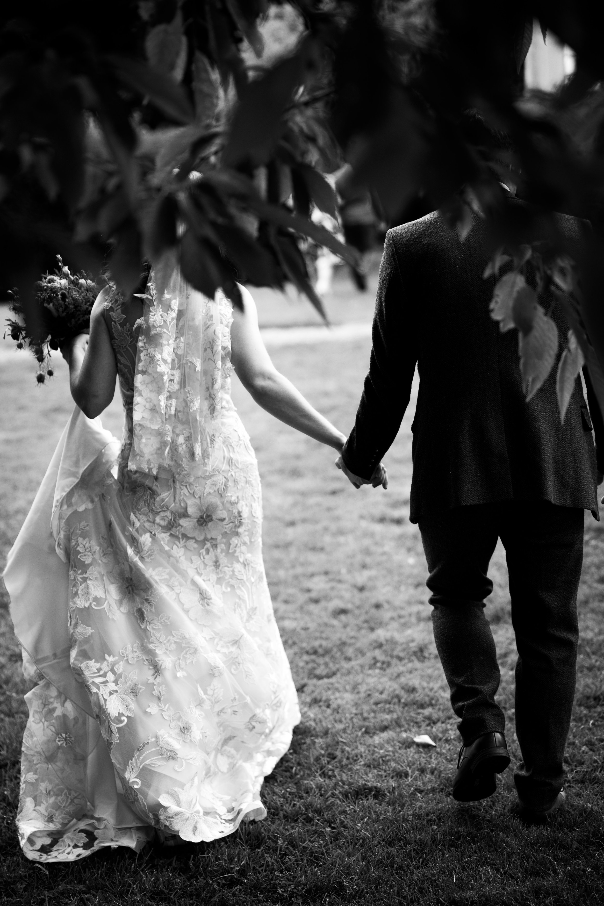 A black and white photo of a couple holding hands outdoors, with the woman wearing a wedding dress and holding a bouquet, and the man dressed in a suit.