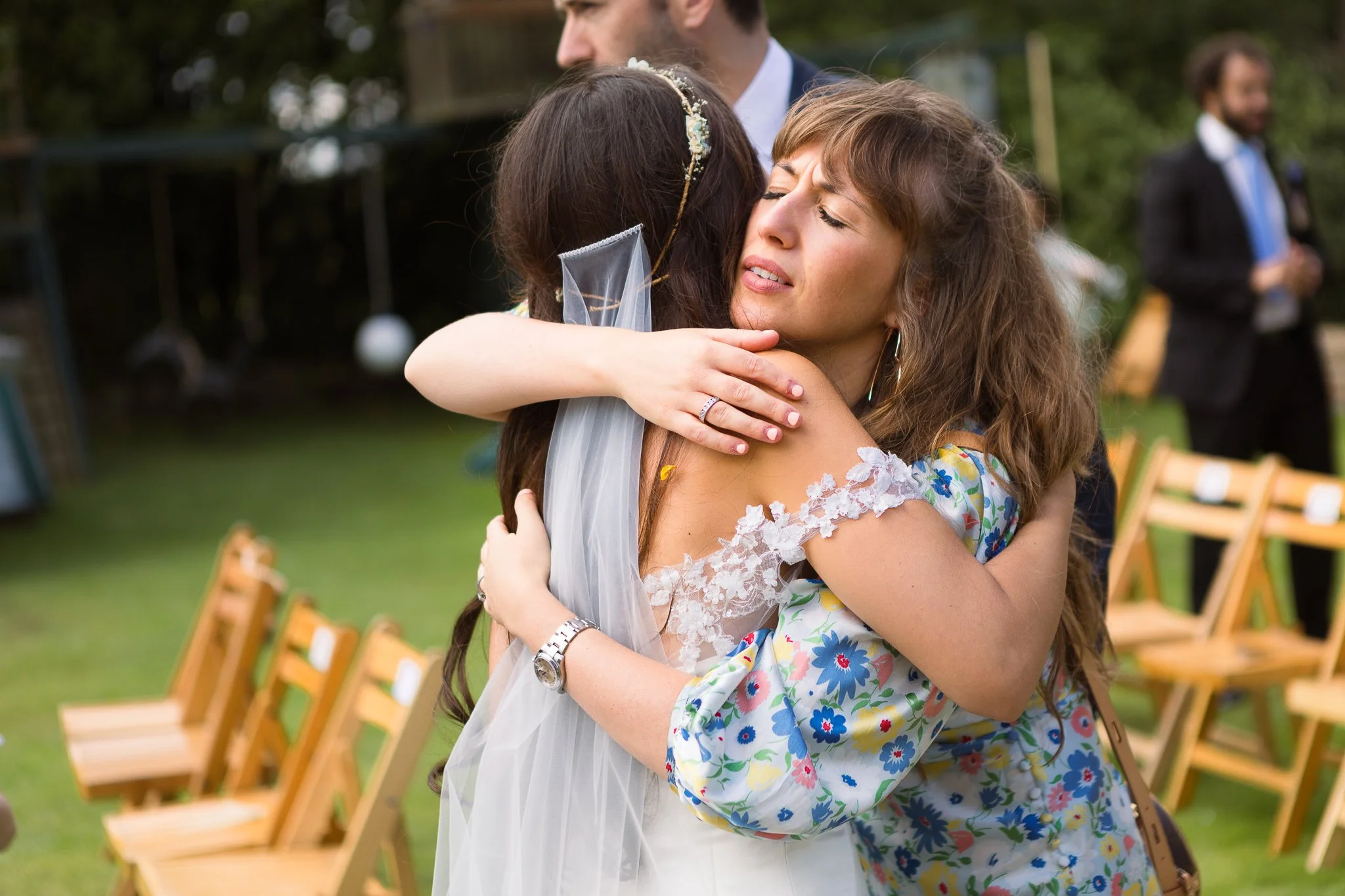 Two women hugging at an outdoor wedding, one dressed in a wedding gown with a veil, the other in a colorful floral dress, with chairs and other wedding guests in the background.