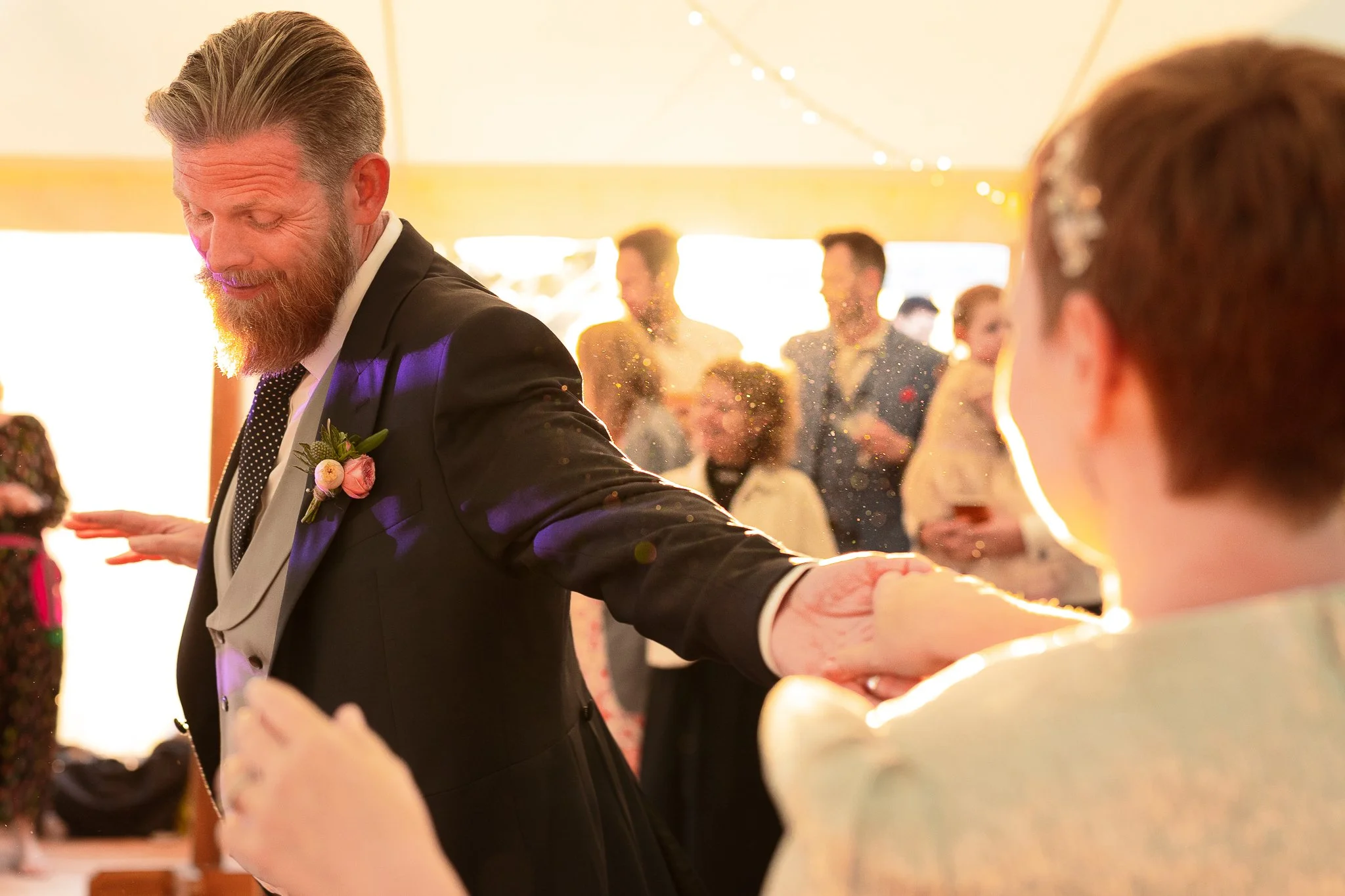 A man with a beard and a boutonniere dances with a woman at a wedding reception, with guests in the background.