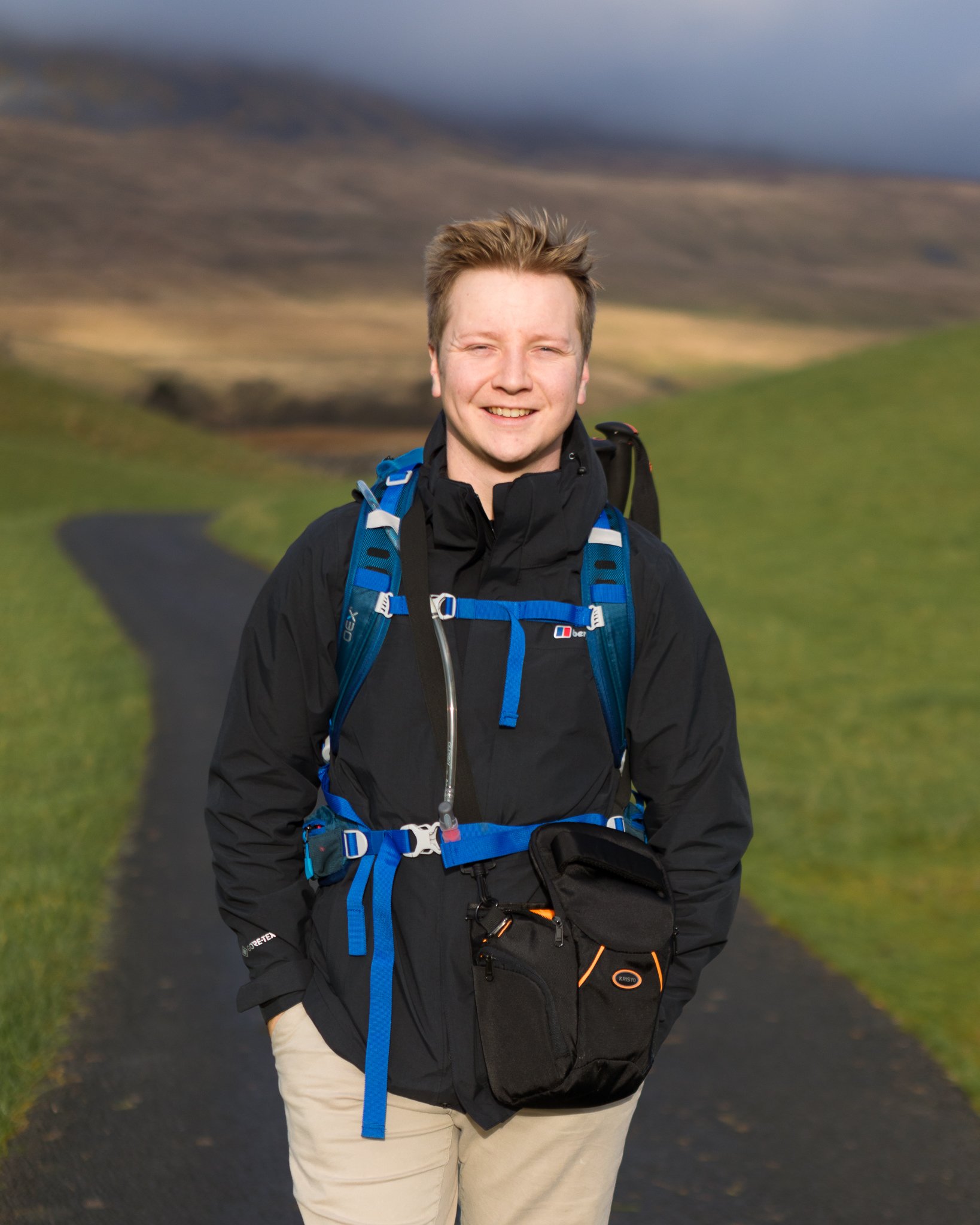 A young man with short, light brown hair smiling outdoors on a cloudy day, wearing a black waterproof jacket, beige pants, and blue hiking gear, standing on a narrow paved path with green grass and rolling hills in the background.