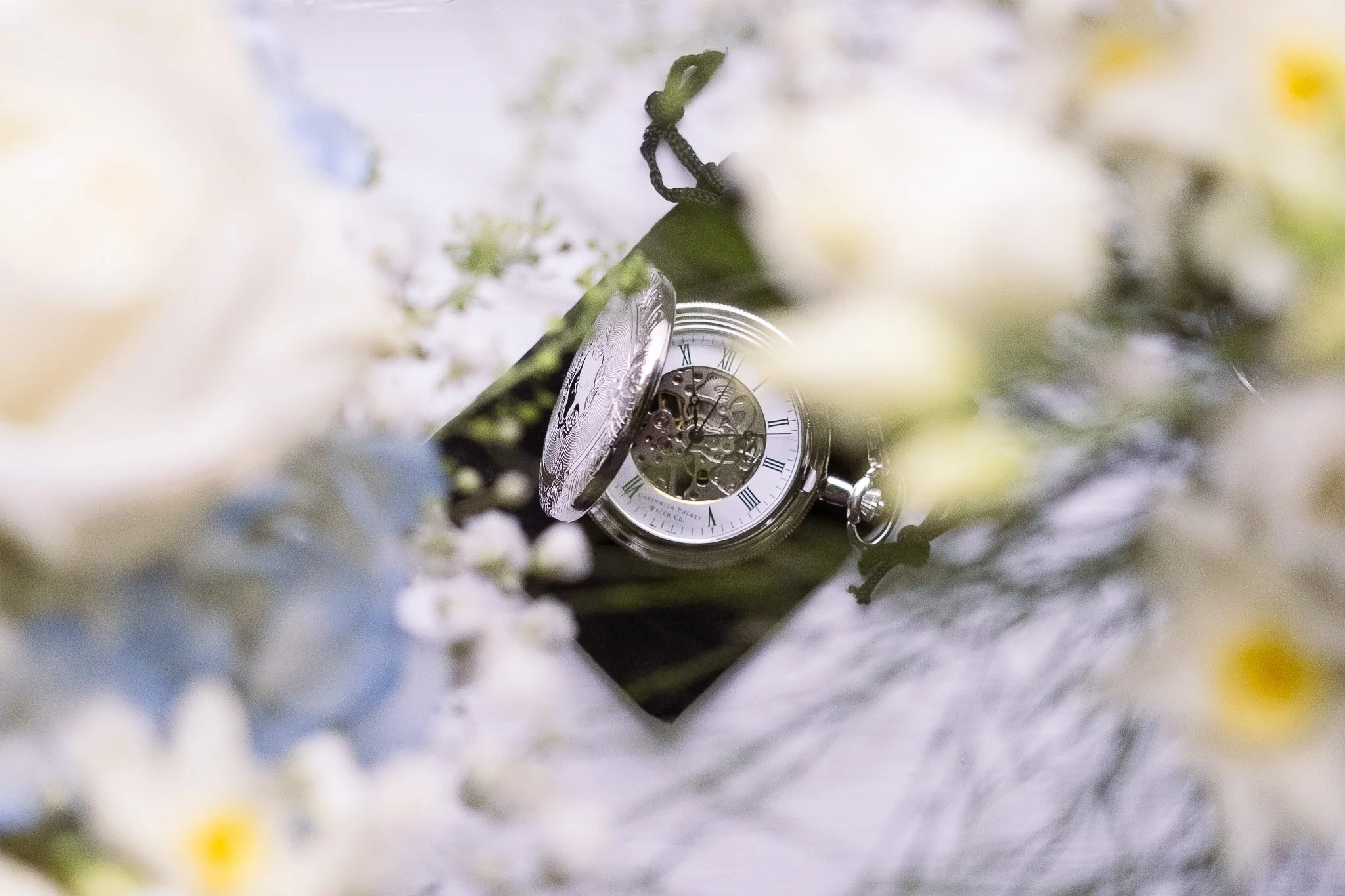 A vintage pocket watch with an open cover showing the clock face and mechanical interior, surrounded by white and blue flowers.