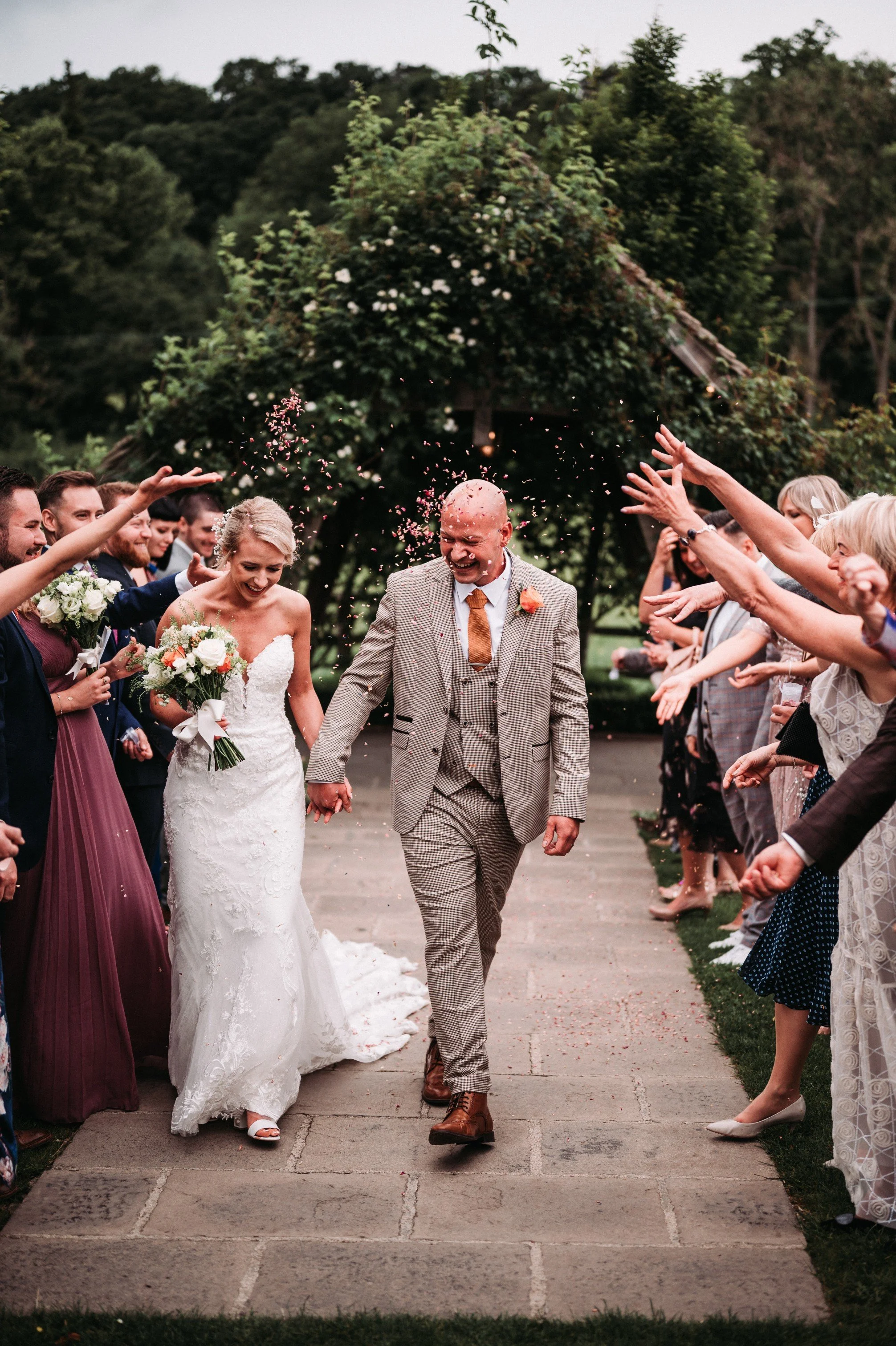 A bride and groom walking hand in hand down an outdoor aisle, surrounded by smiling guests throwing flower petals in celebration.