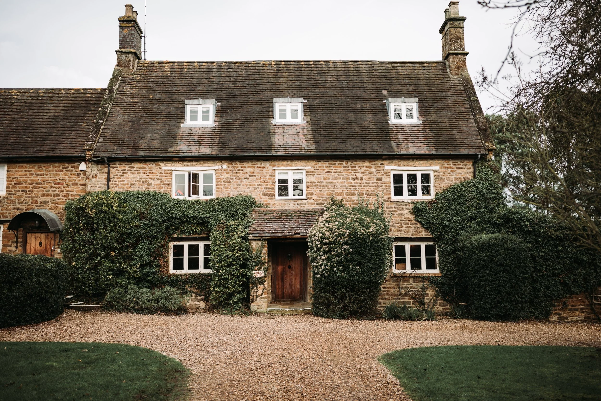 A rustic stone house with a pitched roof, white window frames, and surrounded by lush greenery and shrubs.