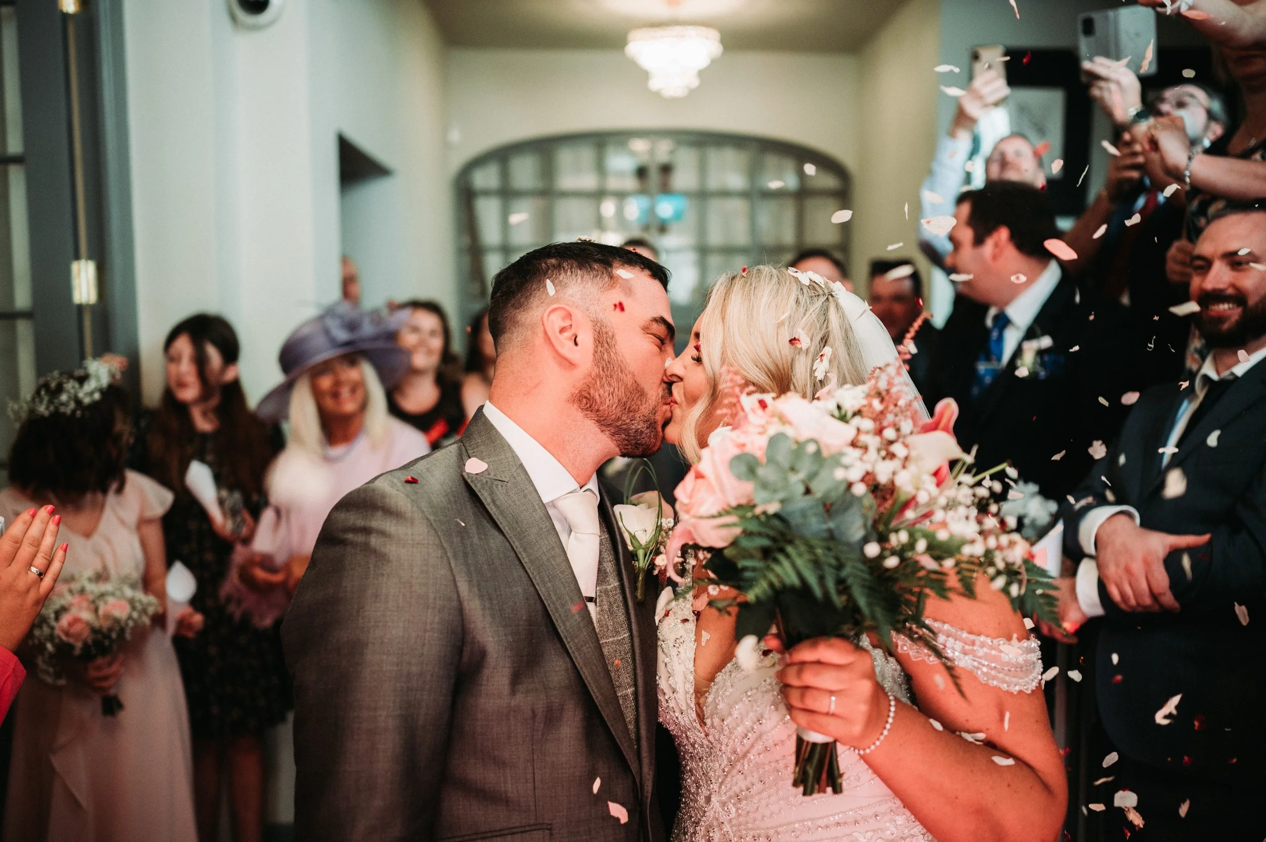 A wedding ceremony where a bride and groom are kissing, surrounded by guests, with confetti falling around them. The bride is holding a bouquet of flowers.