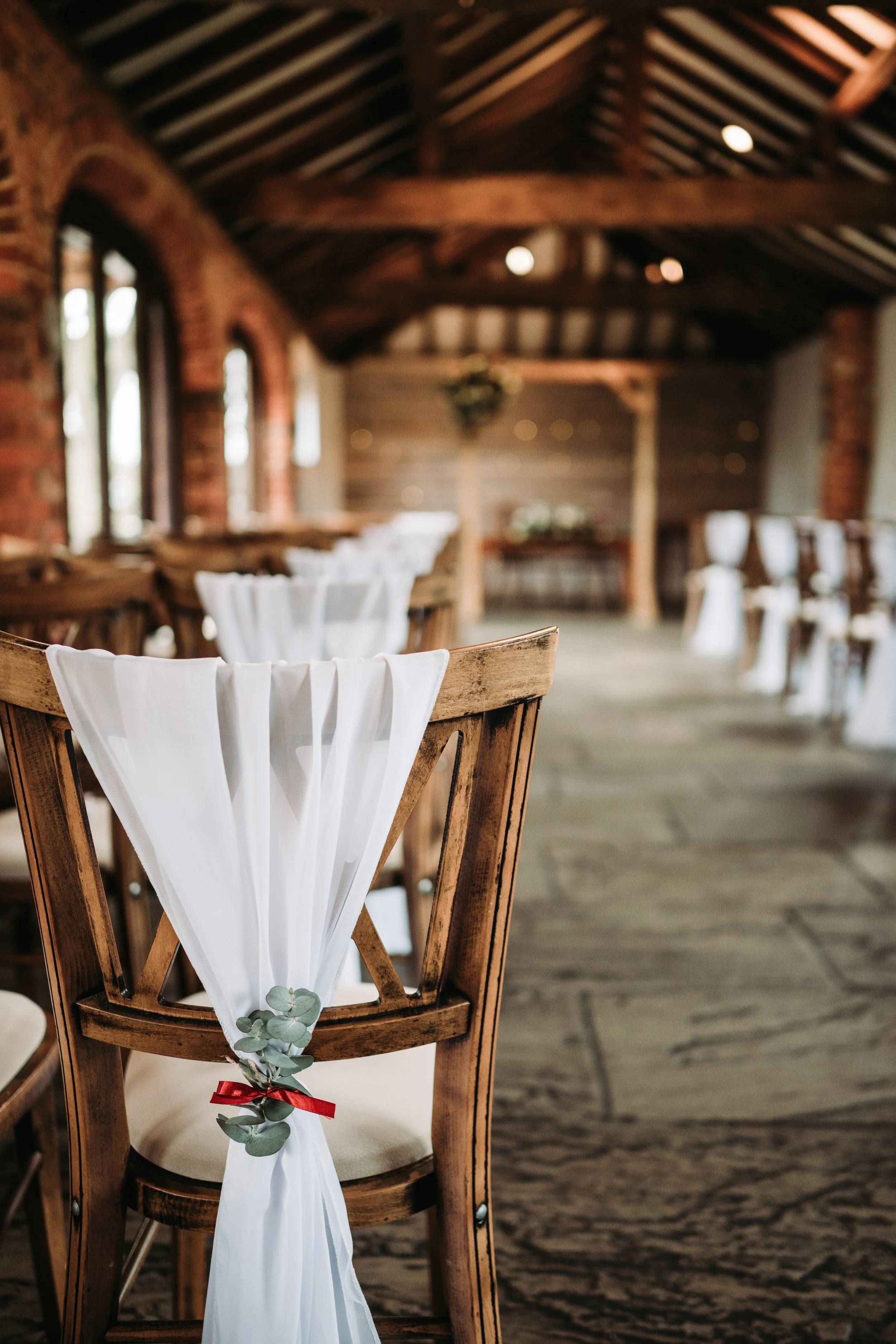 A wooden chair decorated with white fabric, eucalyptus sprigs, and a red ribbon, set up in a rustic venue with exposed brick walls, wooden beams, and large arched windows.