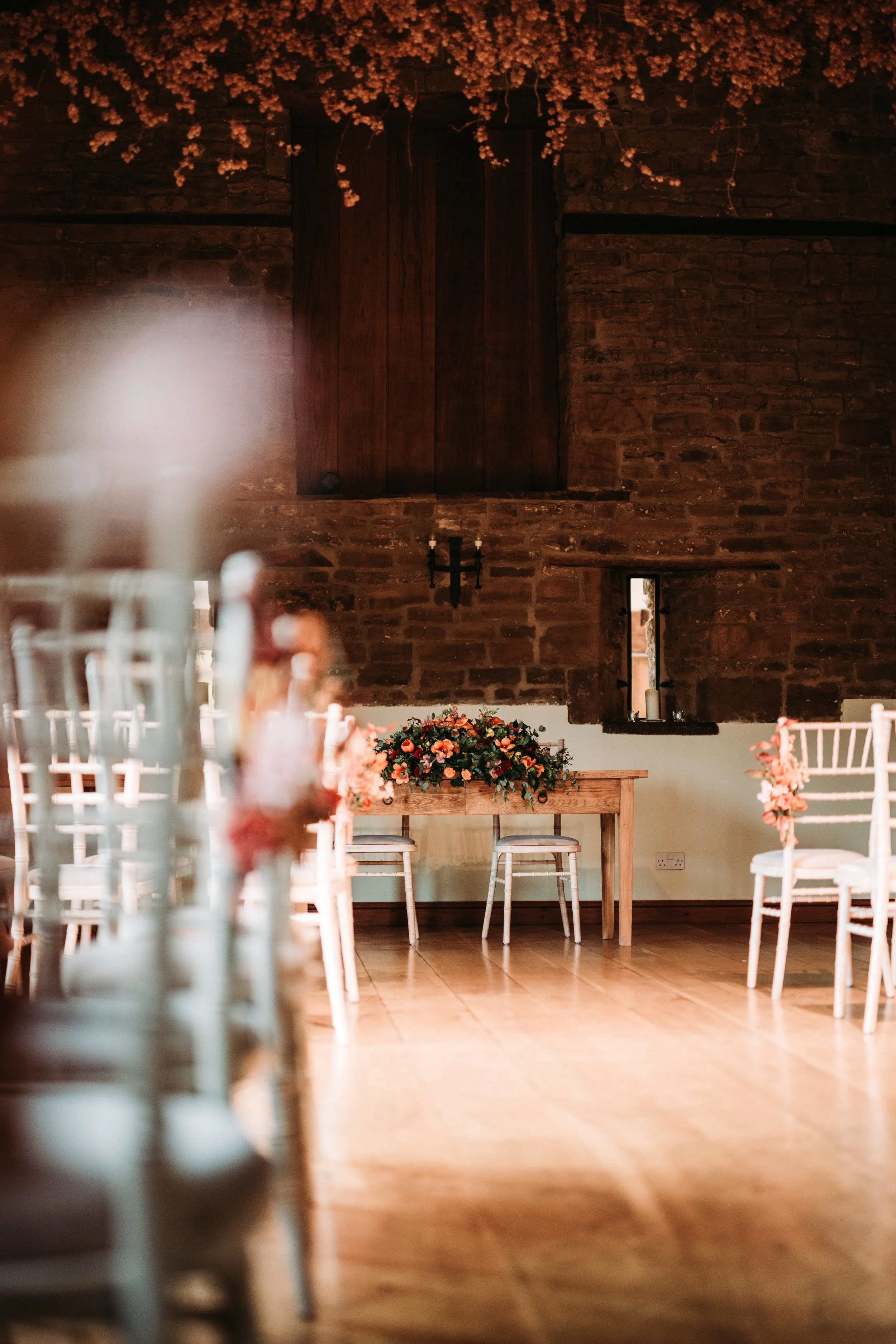 Interior of a rustic event space with wooden beams and brick walls, decorated with floral arrangements and chairs arranged for a gathering.