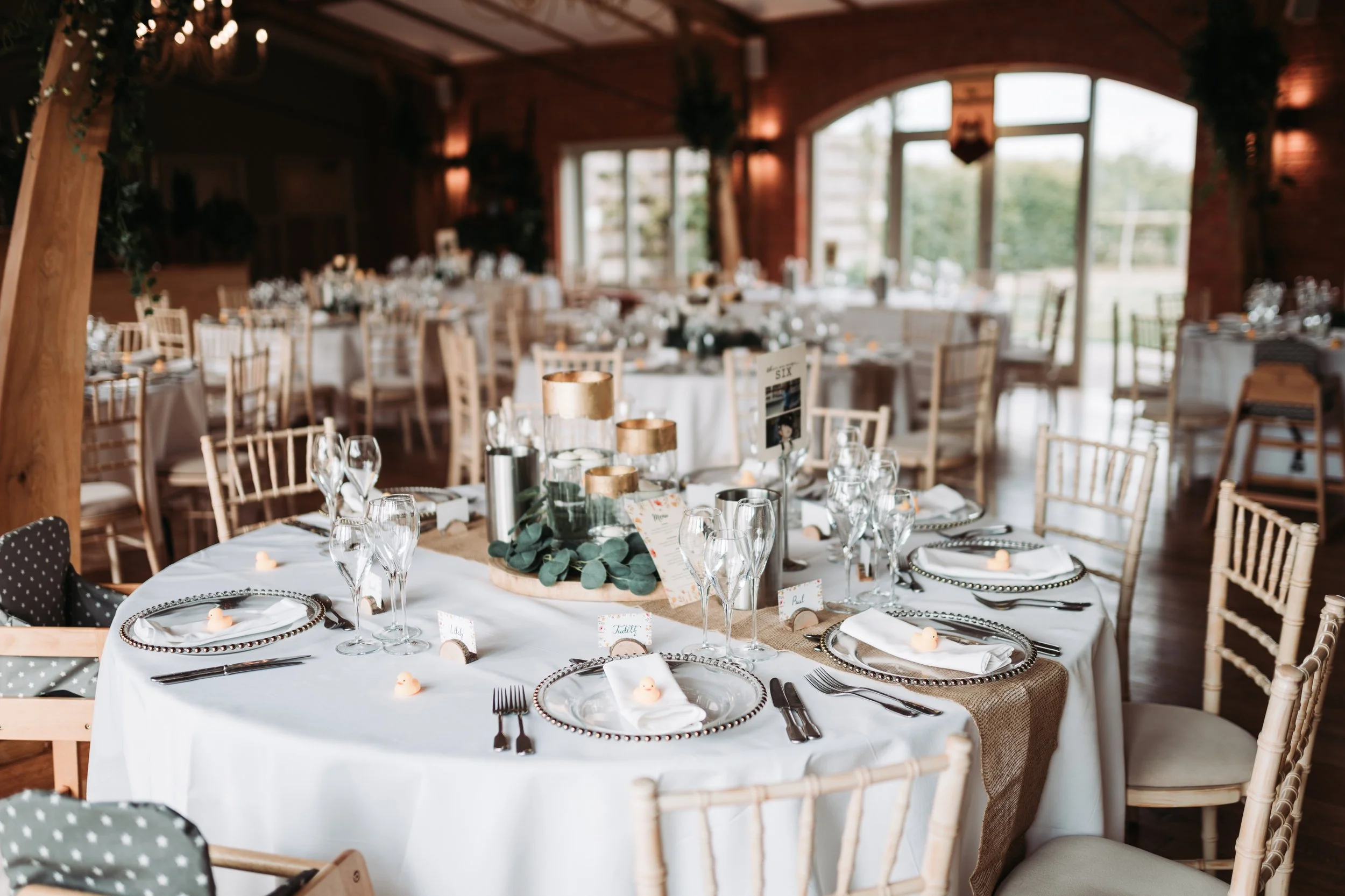 Wedding reception setup with round tables covered in white tablecloths, decorated with candles, greenery, and place settings, in a wooden venue with large windows and natural light.