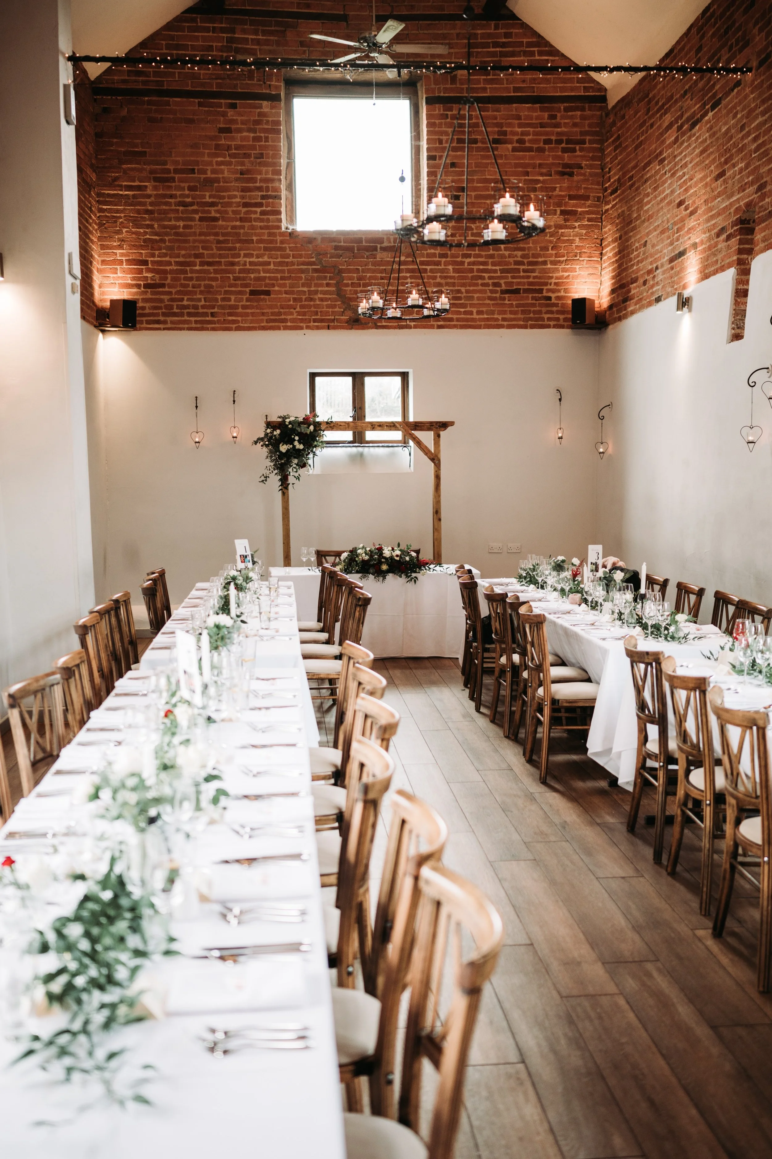 Elegant banquet hall with long reception tables decorated with white tablecloths, centerpieces, and tableware. The room features a high, brick wall ceiling with chandeliers and wall-mounted lights. There is a floral decoration and a wooden arch in the background, indicating a wedding or event setup.