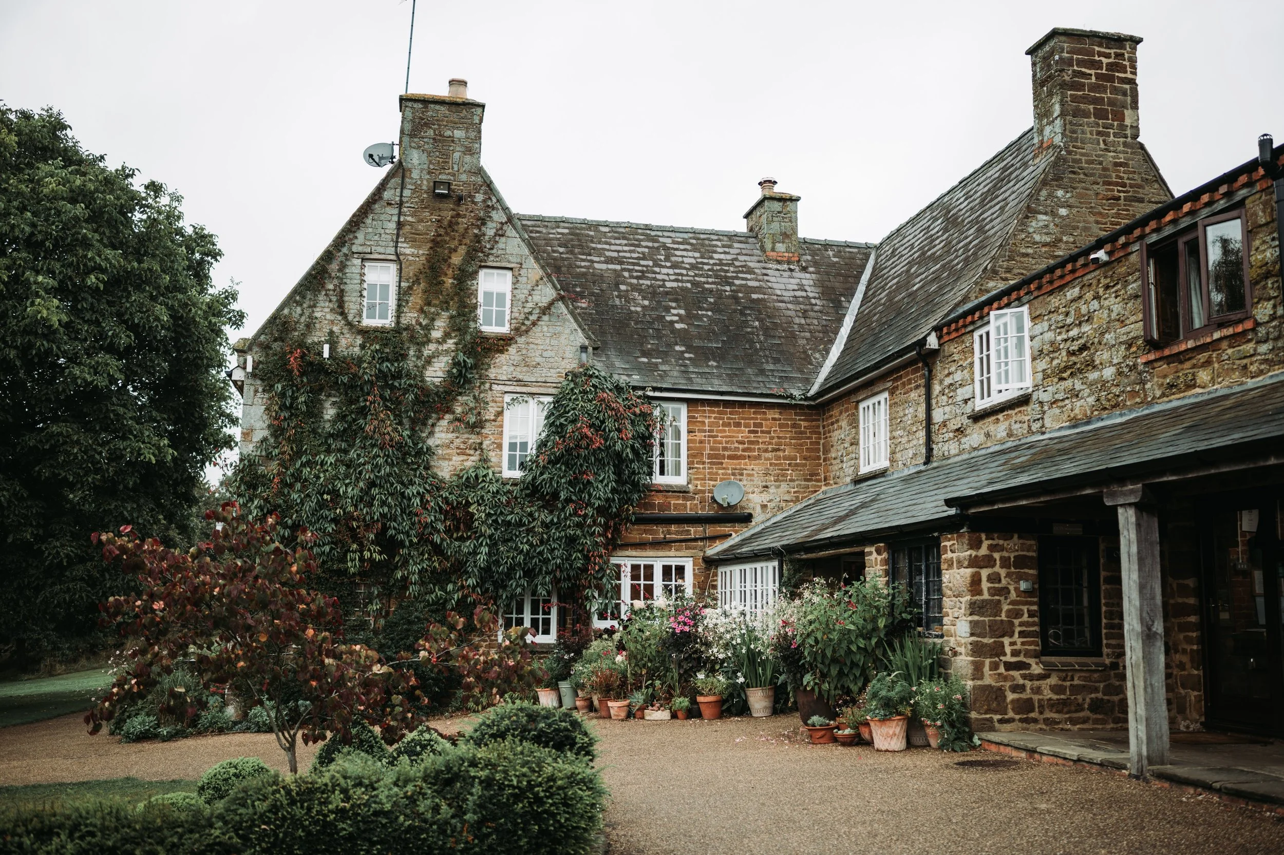A rustic stone house with multiple chimneys, surrounded by a lush garden with potted plants, trees, and flowering bushes.