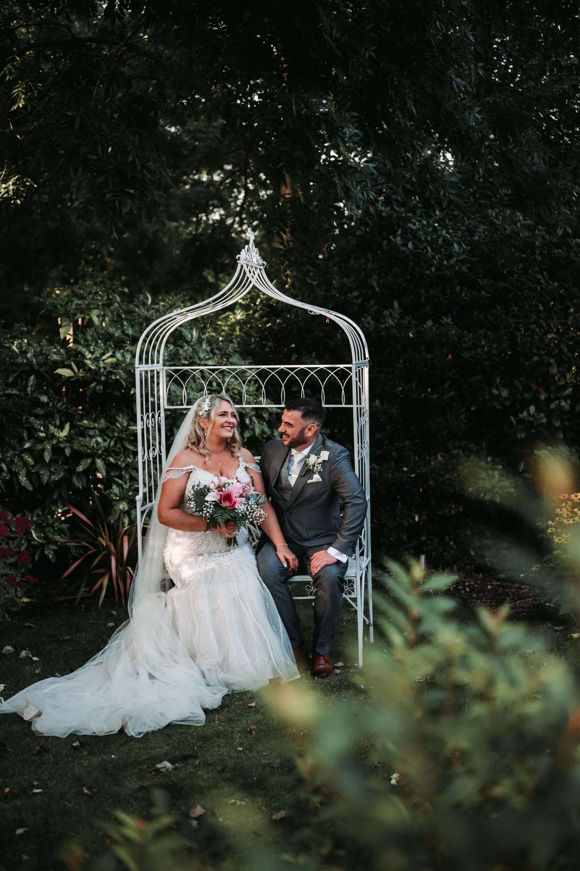 A bride and groom sitting together under a decorative white metal arch in a lush, green outdoor setting during their wedding ceremony. The bride is dressed in a white wedding gown holding a bouquet of pink and white flowers, and the groom is in a grey suit.