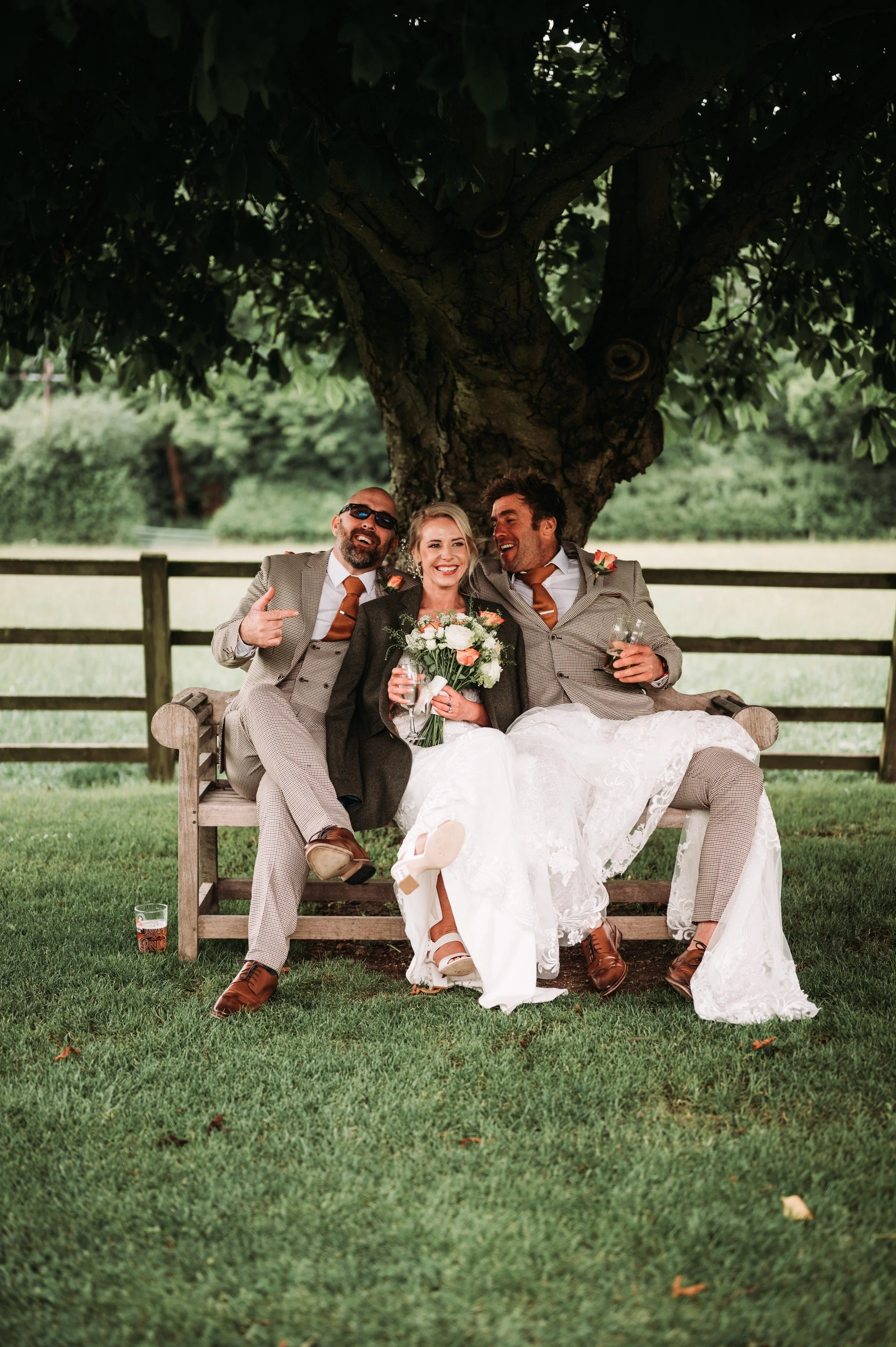 Three people are sitting on a bench under a large tree in a park, smiling and celebrating a wedding. The woman in the center is holding a bouquet, wearing a white dress, and flanked by two men in suits, one of whom is pointing and the other holding a glass of drink.