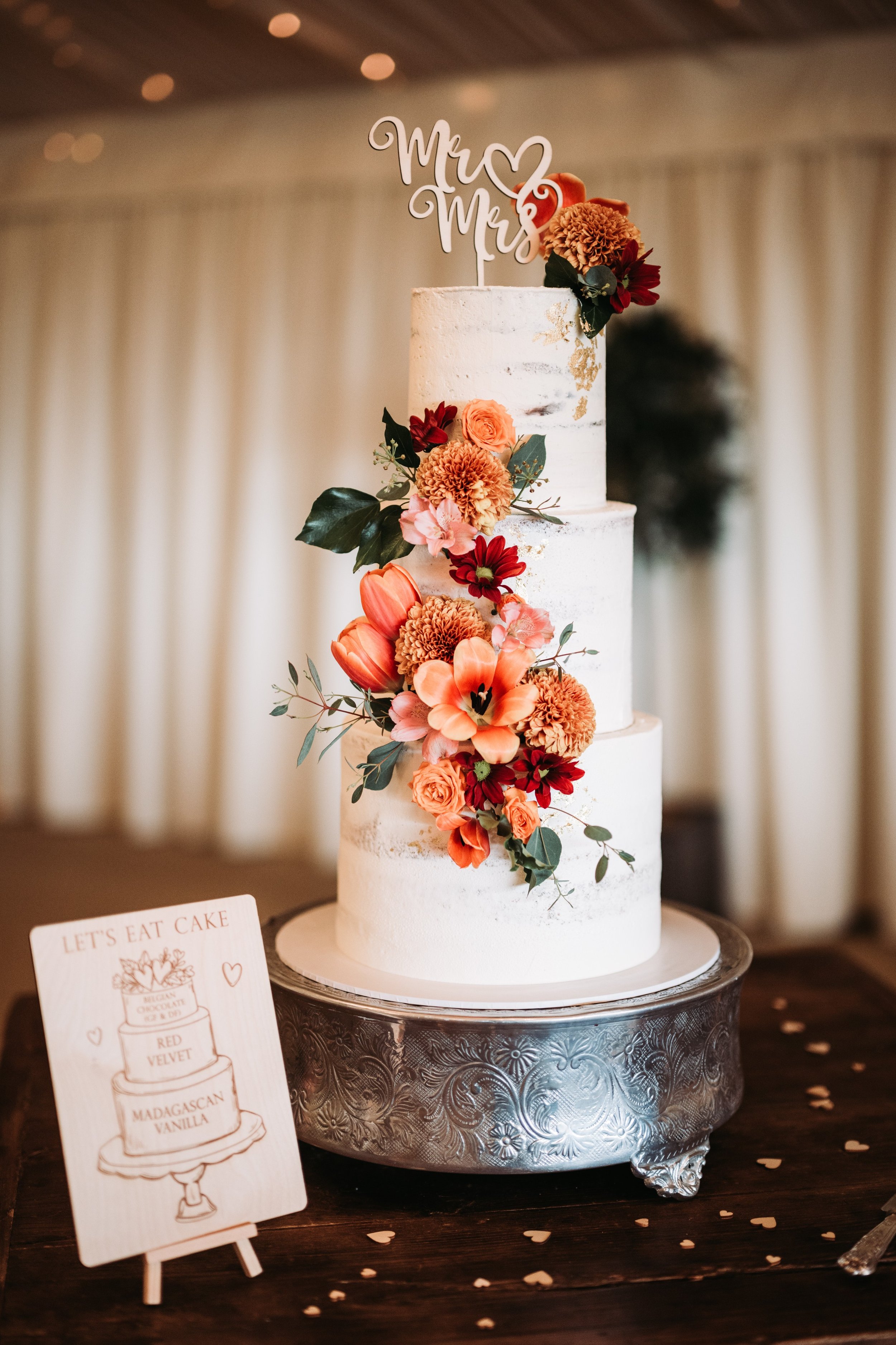 A tall, white wedding cake decorated with orange and red flowers and greenery, topped with a 'Mr & Mrs' cake topper, on an ornate silver cake stand on a dark wooden table with scattered paper hearts.