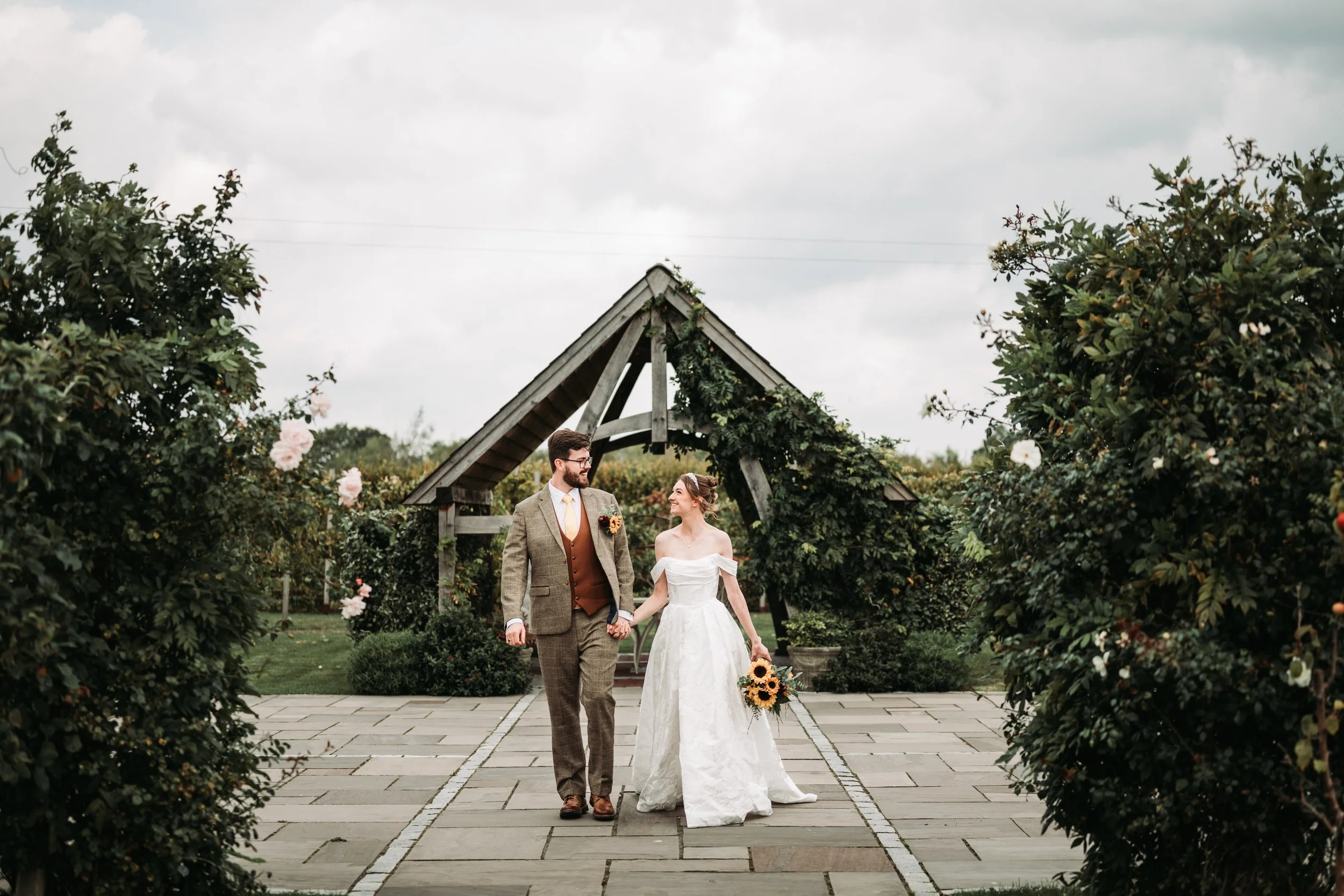 A bride and groom holding hands, walking outdoors in a garden setting with lush bushes and flowers, under a cloudy sky, during a wedding ceremony.