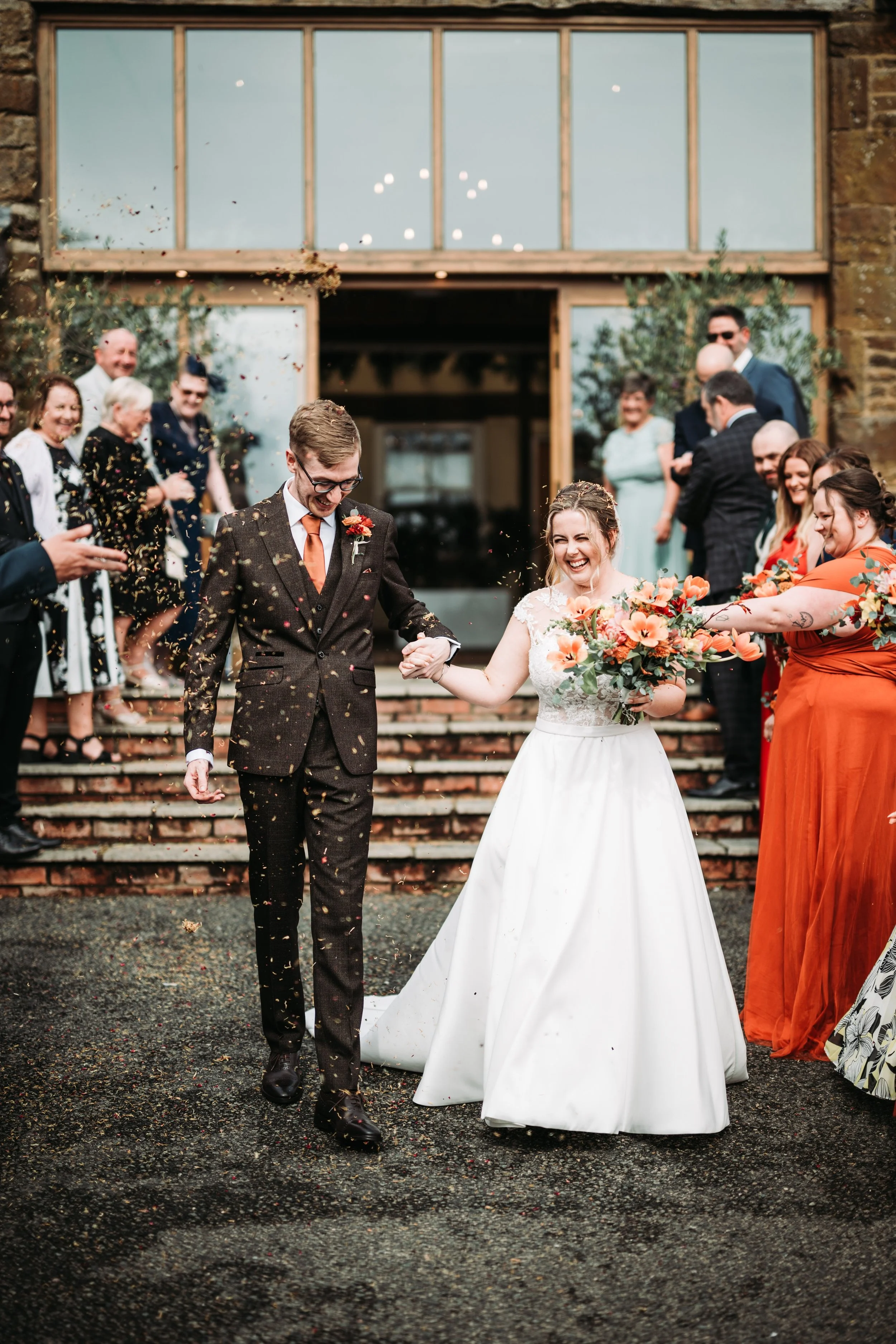 A bride and groom walking down steps outside a building after their wedding, smiling and holding hands, surrounded by friends and family throwing confetti.
