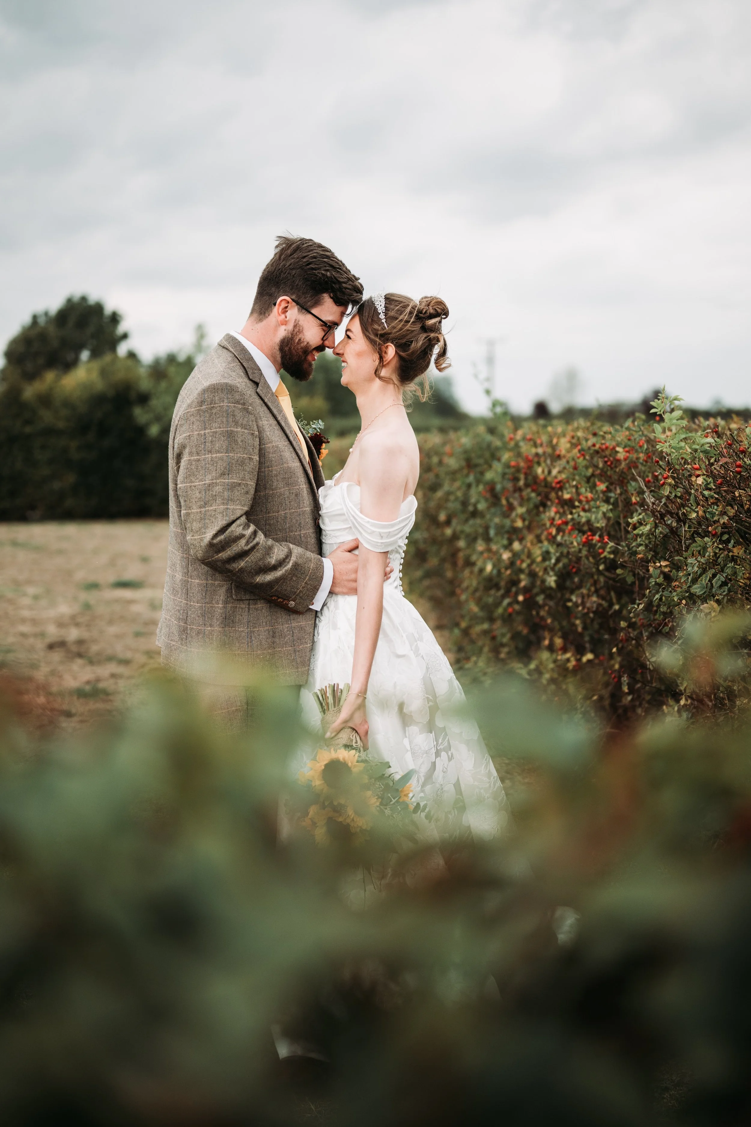 A couple in wedding attire standing outdoors in a field with bushes and overcast sky, about to kiss with foreheads touching.