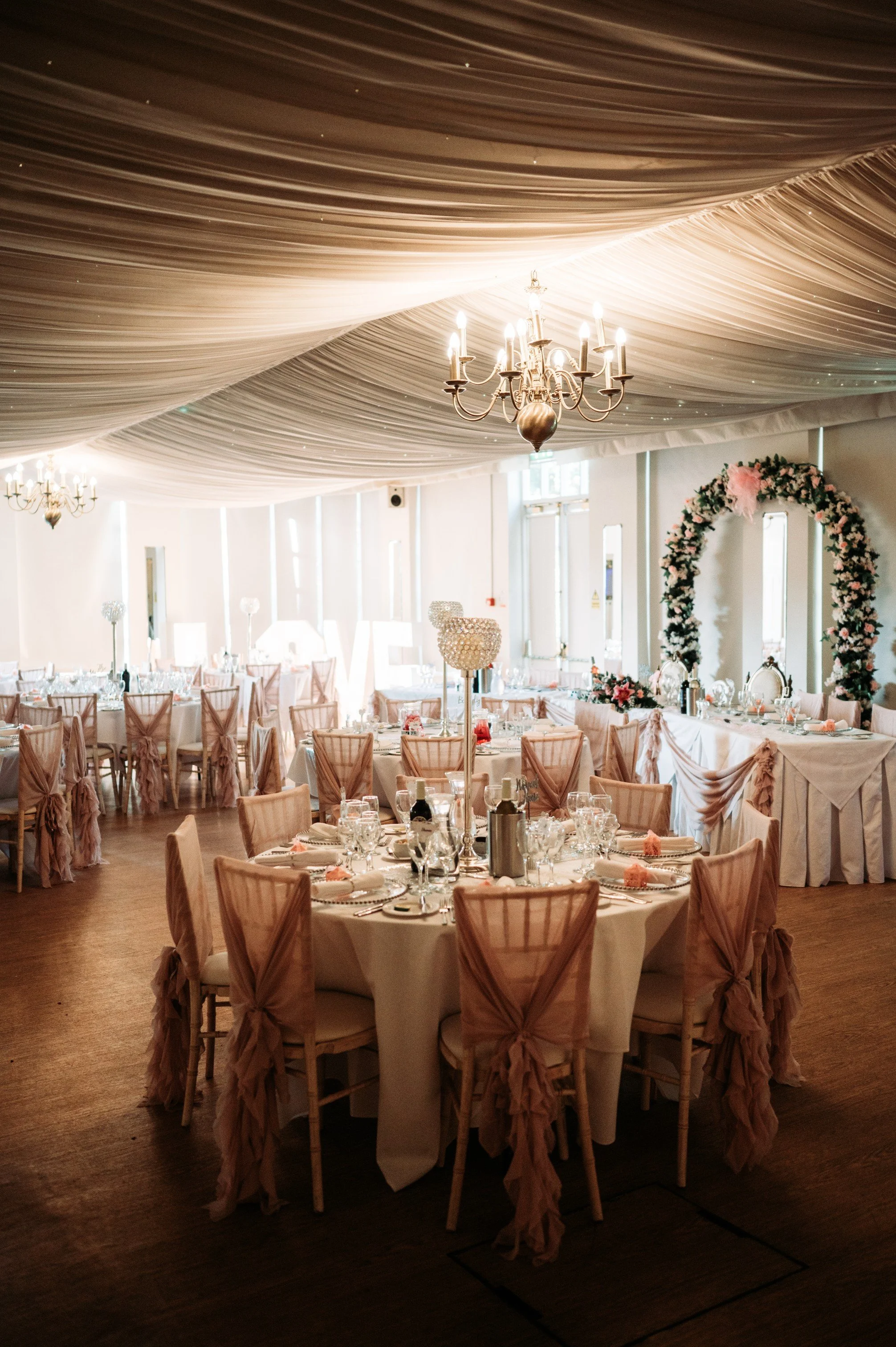 A decorated event hall with round tables covered in white tablecloths, pink chairs with bows, floral centerpieces, and an altar arch adorned with pink and white flowers. The ceiling has draped fabric with small lights, and there are chandeliers hanging from above.