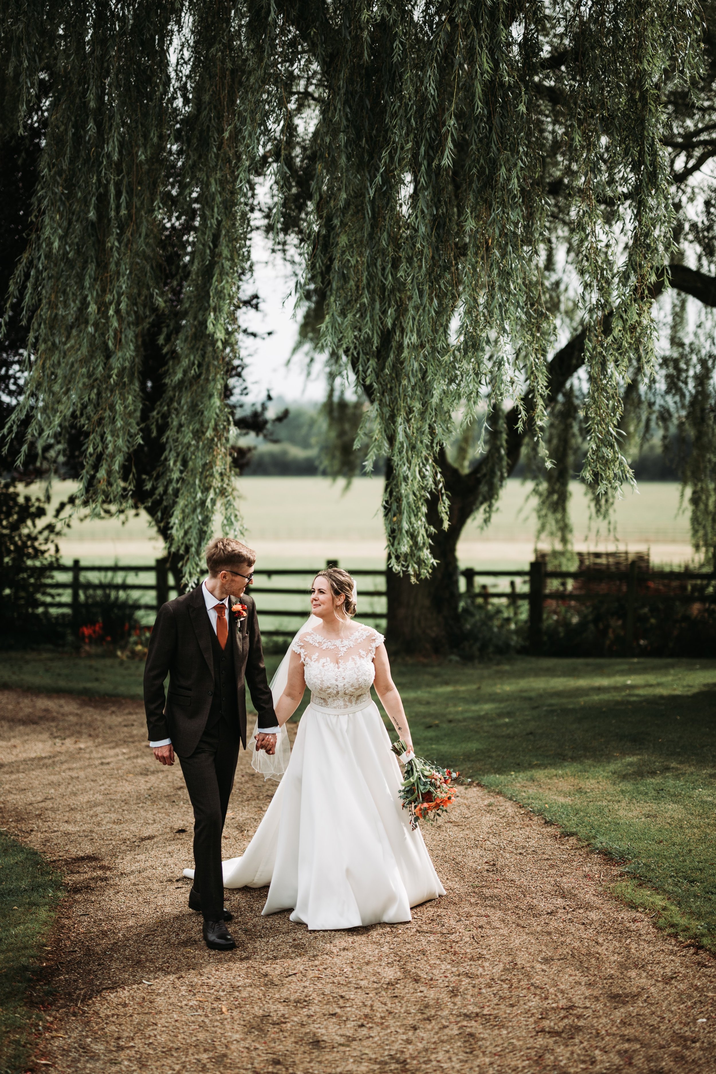 A bride and groom holding hands and walking on a pathway outdoors, with lush green grass and trees in the background, under a large hanging tree.