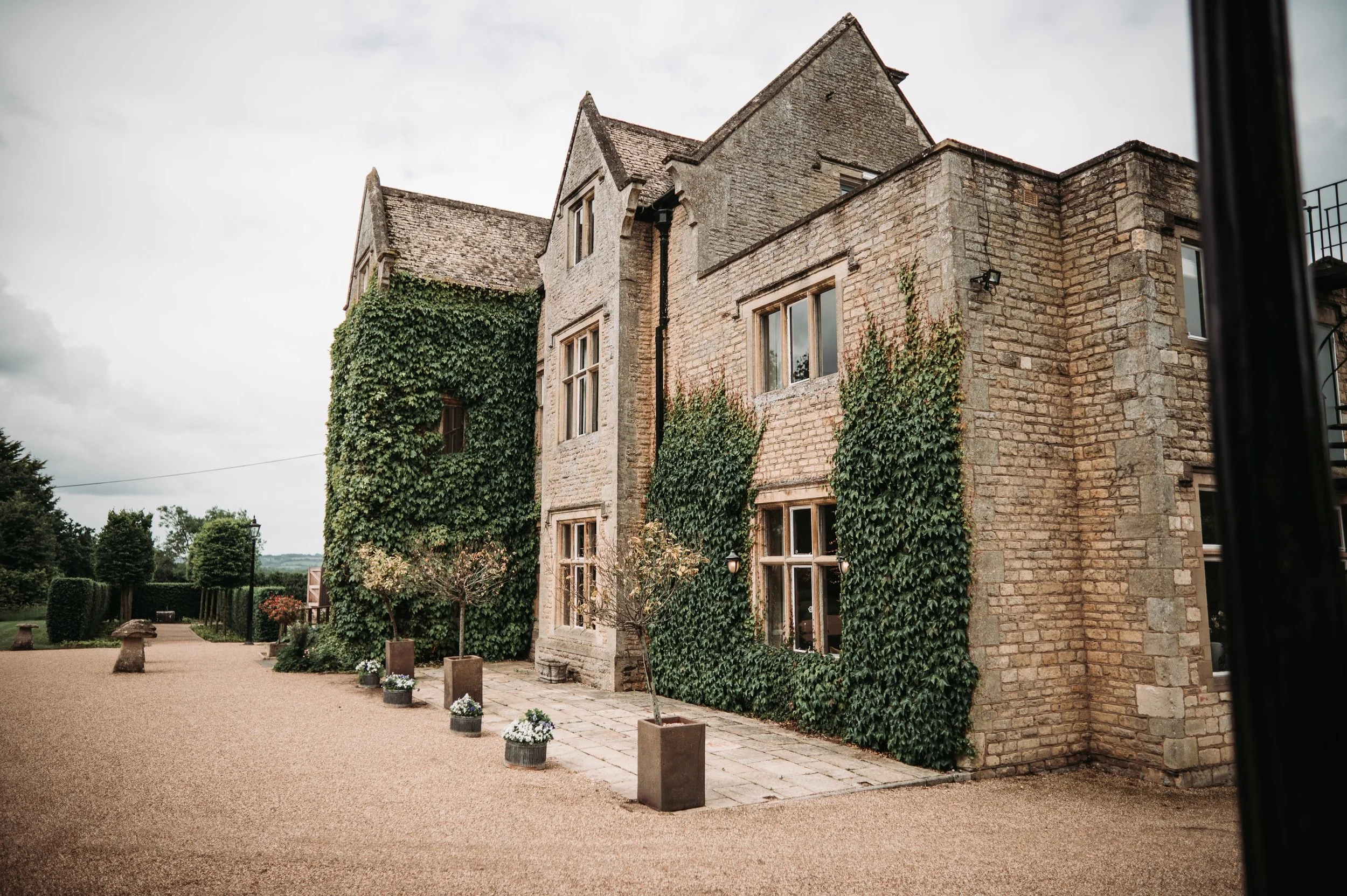 A historic stone building with ivy growing on the walls, surrounded by a gravel pathway and small potted plants, under a cloudy sky.