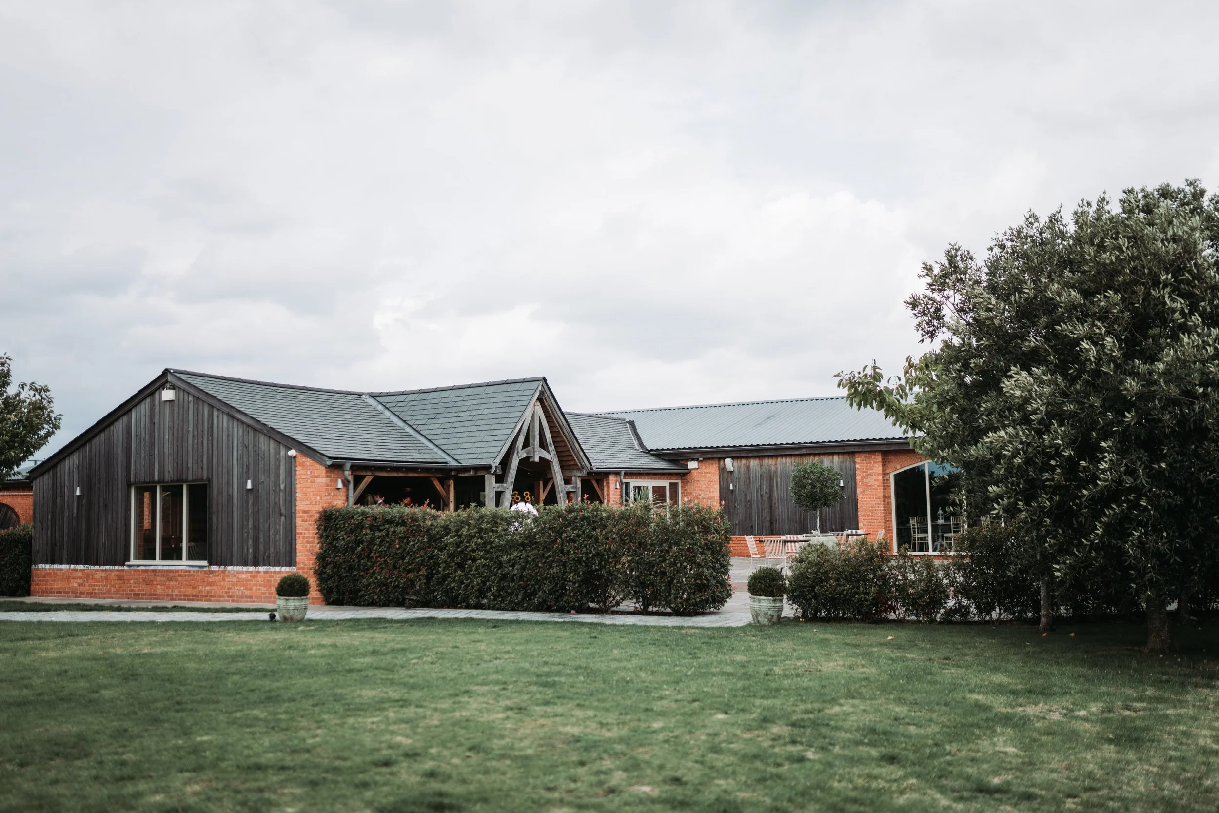 A large backyard with a sprawling, well-maintained green lawn, surrounded by trees and shrubs. In the background, there is a modern barn-style building with a combination of brick and dark wood siding, and a gabled roof. The sky is overcast with gray clouds.