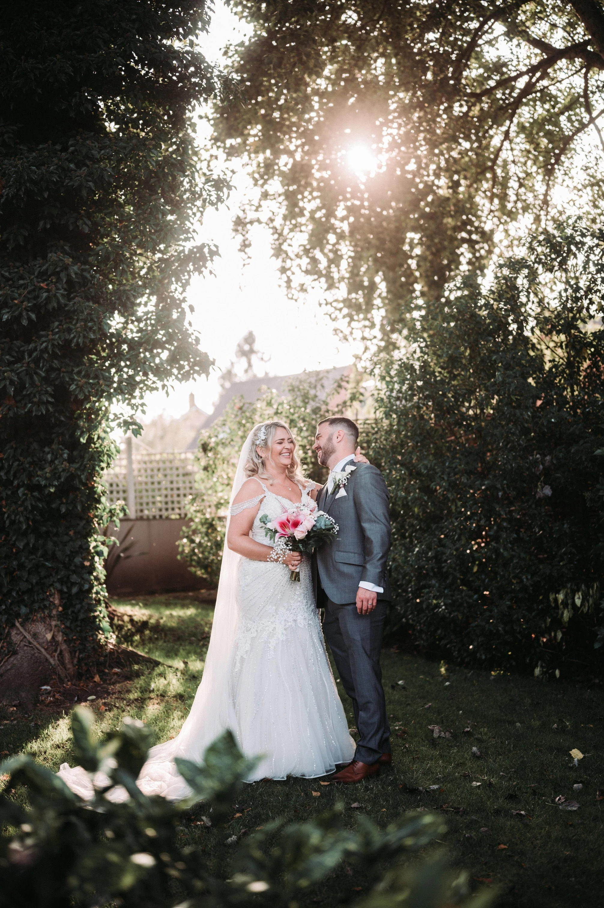 A bride and groom standing together outdoors during their wedding ceremony, surrounded by trees and greenery with sunlight shining through the branches.