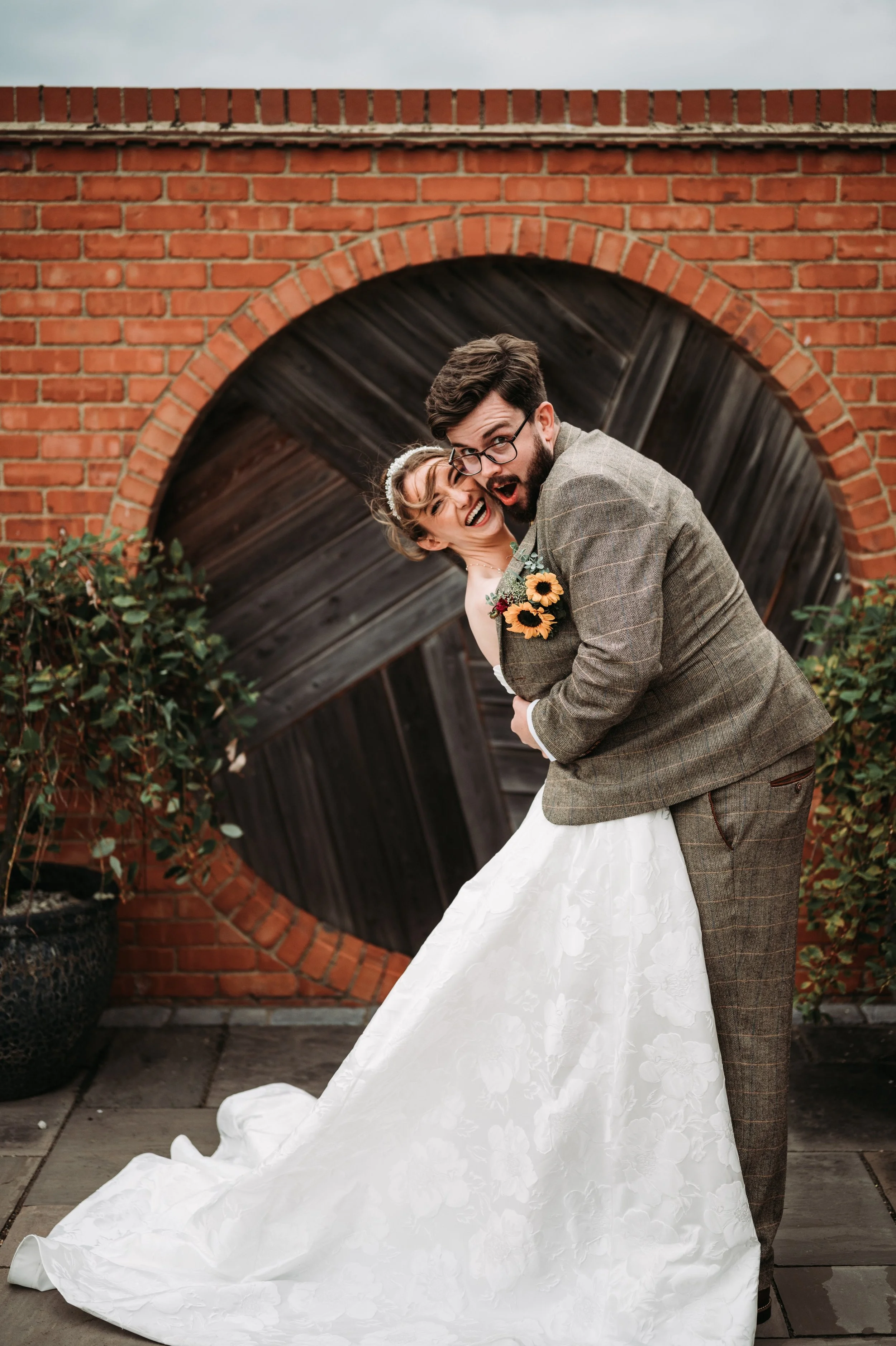 A bride and groom are smiling and posing playfully in front of a brick wall with a circular opening, with the bride wearing a white floral gown and a small veil, and the groom wearing a plaid suit with a sunflower boutonniere.