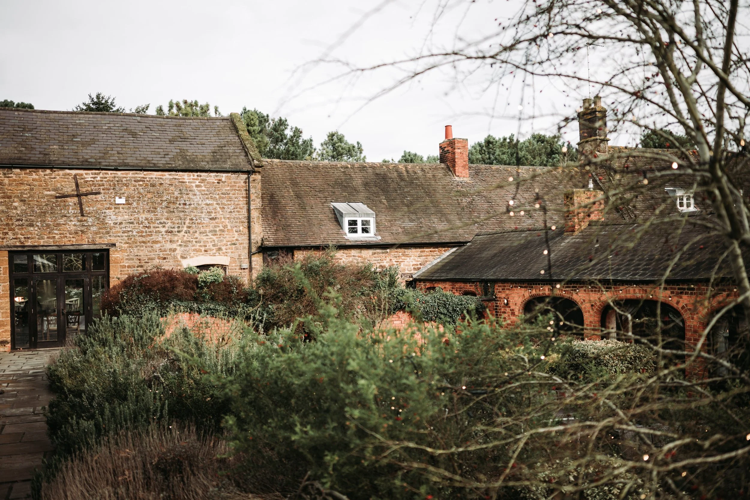 Exterior of a brick house with a sloped roof, dormer windows, and chimneys, surrounded by trees and shrubs, with the sky overcast.