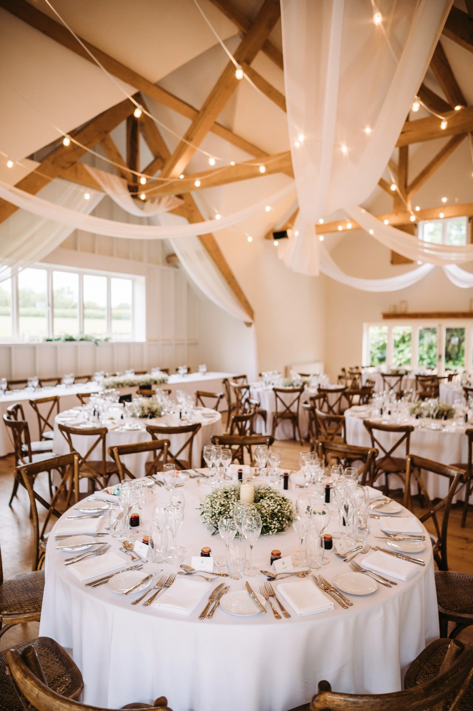 Elegant event space with round tables covered in white tablecloths, set with glassware, plates, and silverware. Centerpieces of baby's breath flowers and candles adorn each table. The ceiling features exposed wooden beams, sheer drapes, and string lights, creating a warm ambiance. Large windows allow natural light into the room.