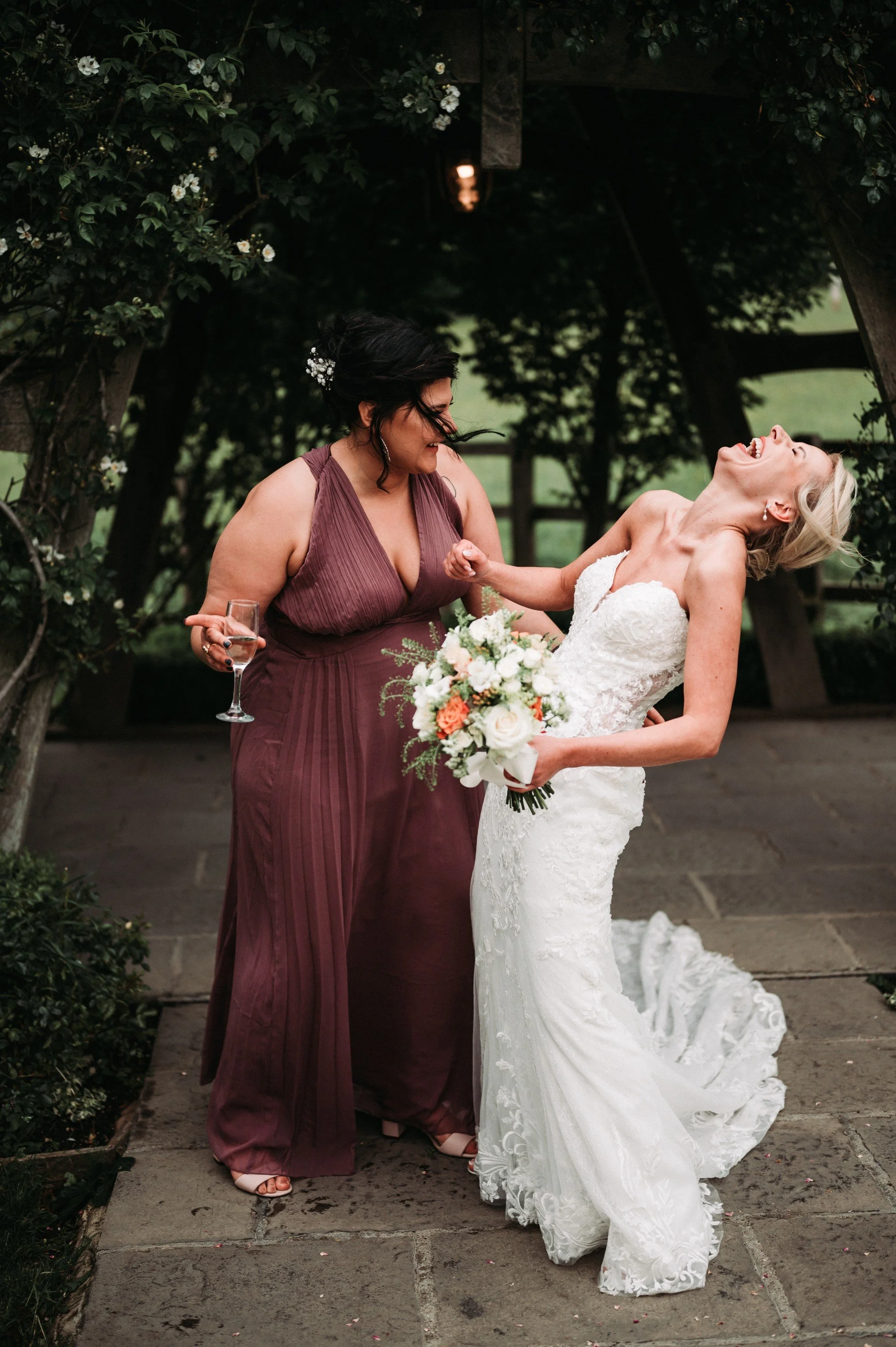 Two women, one in a white wedding dress and the other in a burgundy dress, laugh together outdoors under a wooden arch decorated with flowers. The bride holds a bouquet, and the other woman has a glass of champagne.
