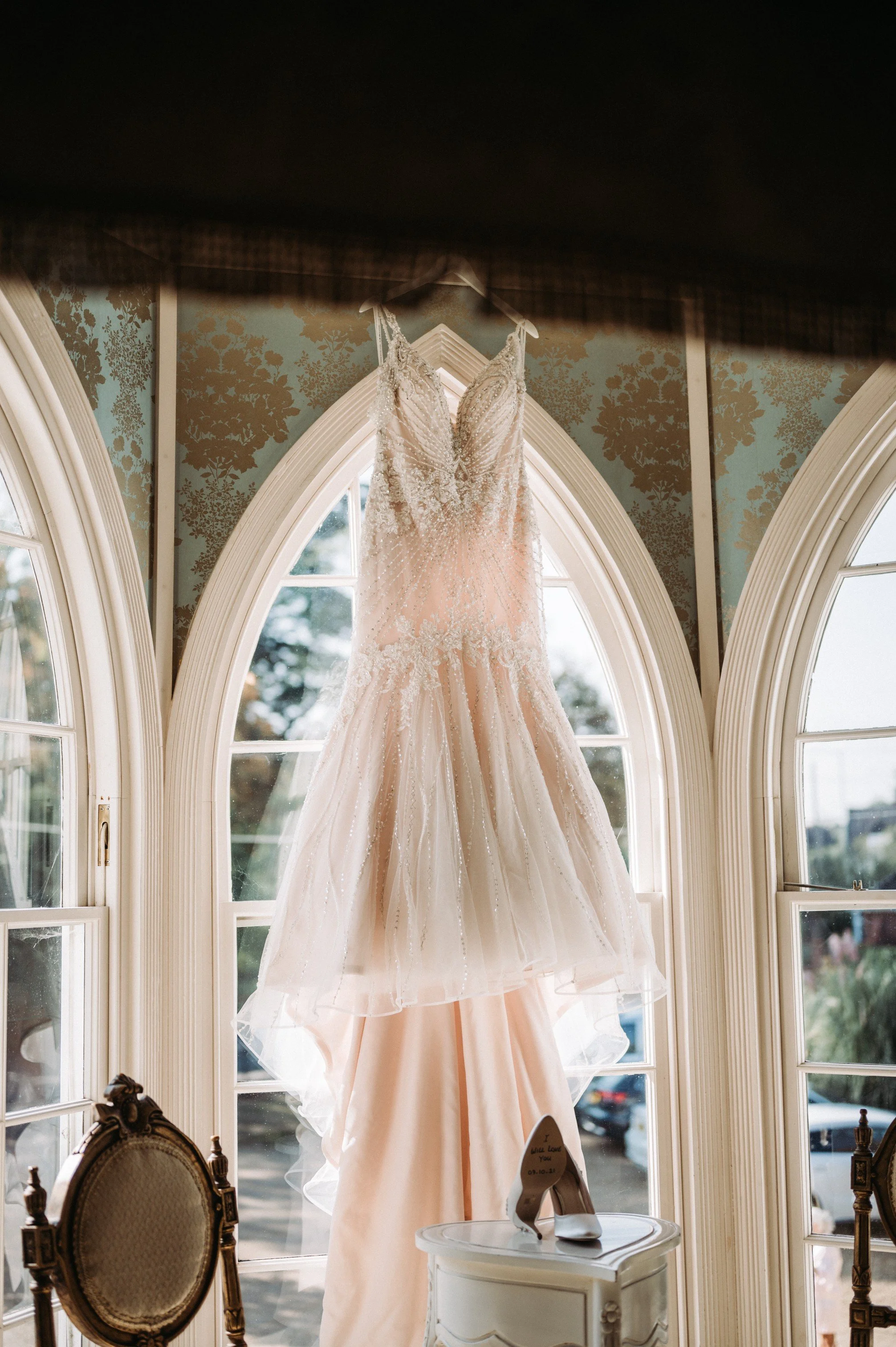 A wedding dress hanging in front of large arched windows with stained glass details, sunlight streaming in, and a pair of white high heels on a small table below.