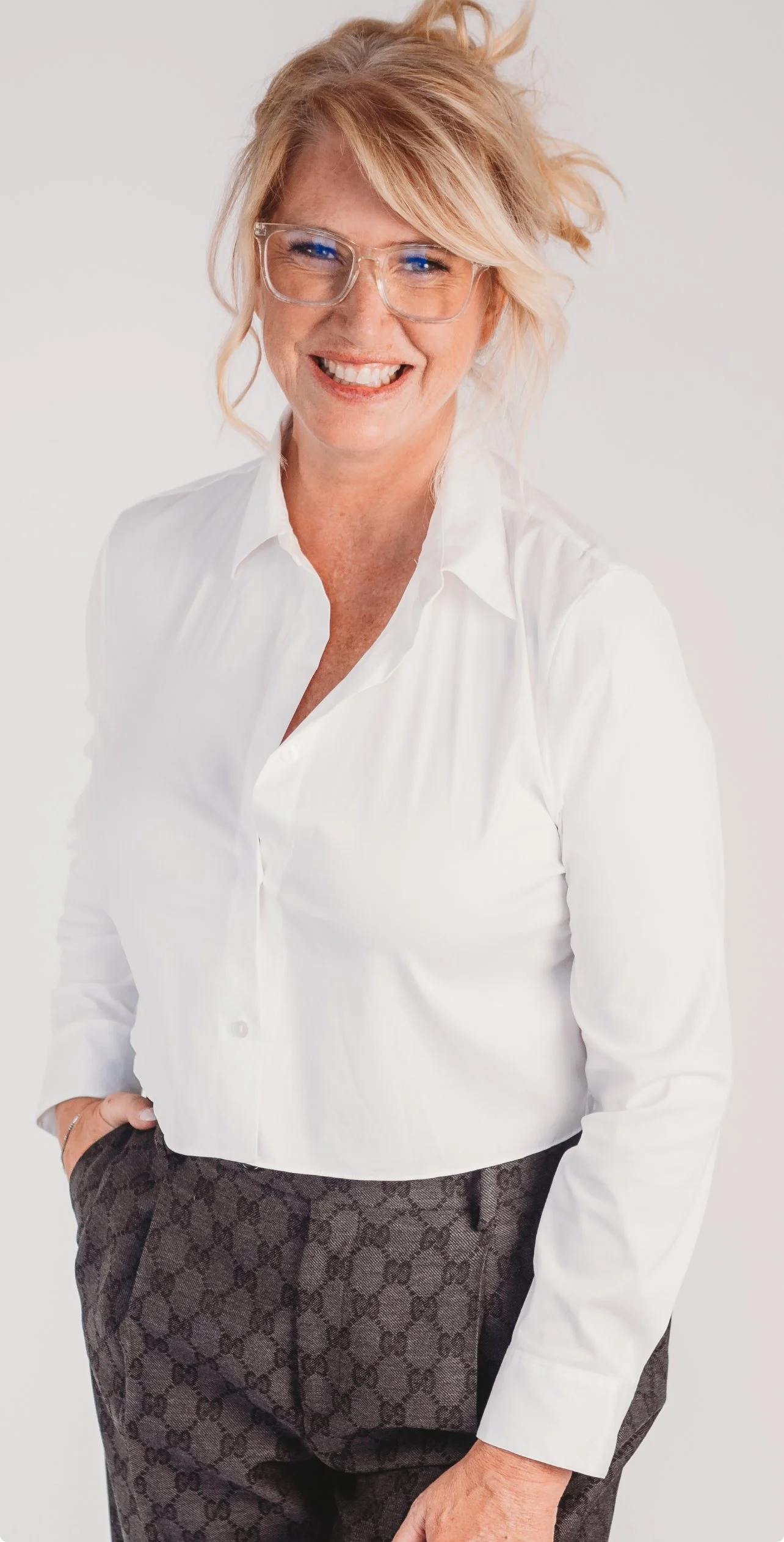 A smiling woman with blonde hair and glasses, wearing a white blouse and patterned pants, standing against a plain white background.