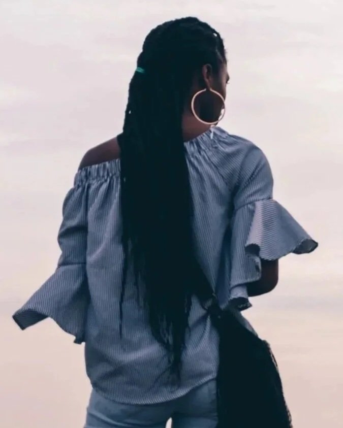 A woman with long dreadlocks wearing large hoop earrings and a blue striped off-shoulder ruffled top, standing outdoors against cloudy sky, representing strength and confidence.