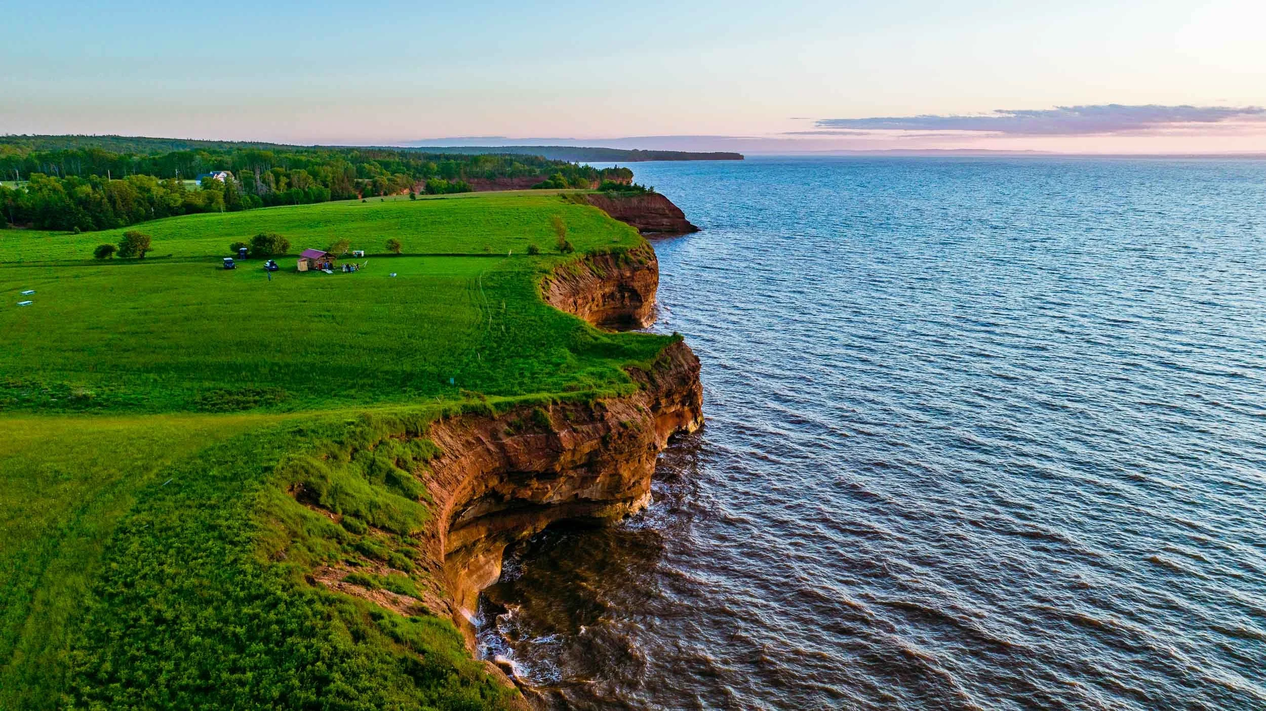 Aerial view of green farmland with scattered trees and small structures along a cliff overlooking the ocean during sunset.