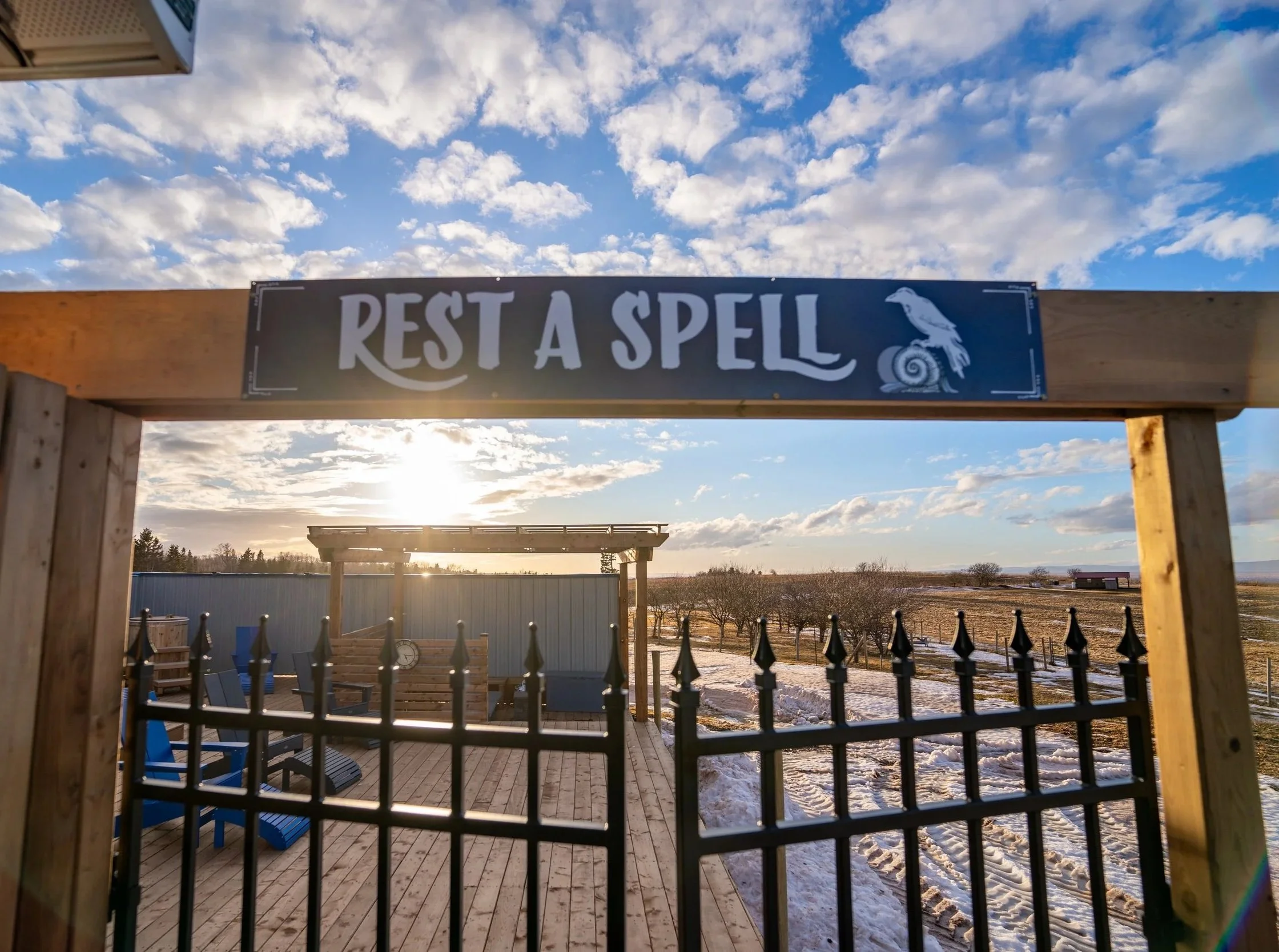 A wooden entrance gate with a sign that reads 'Rest a Spell' and a bird on a snail, overlooking a deck with outdoor seating, a fence, snow-covered ground, and a rural landscape under a partly cloudy sky.