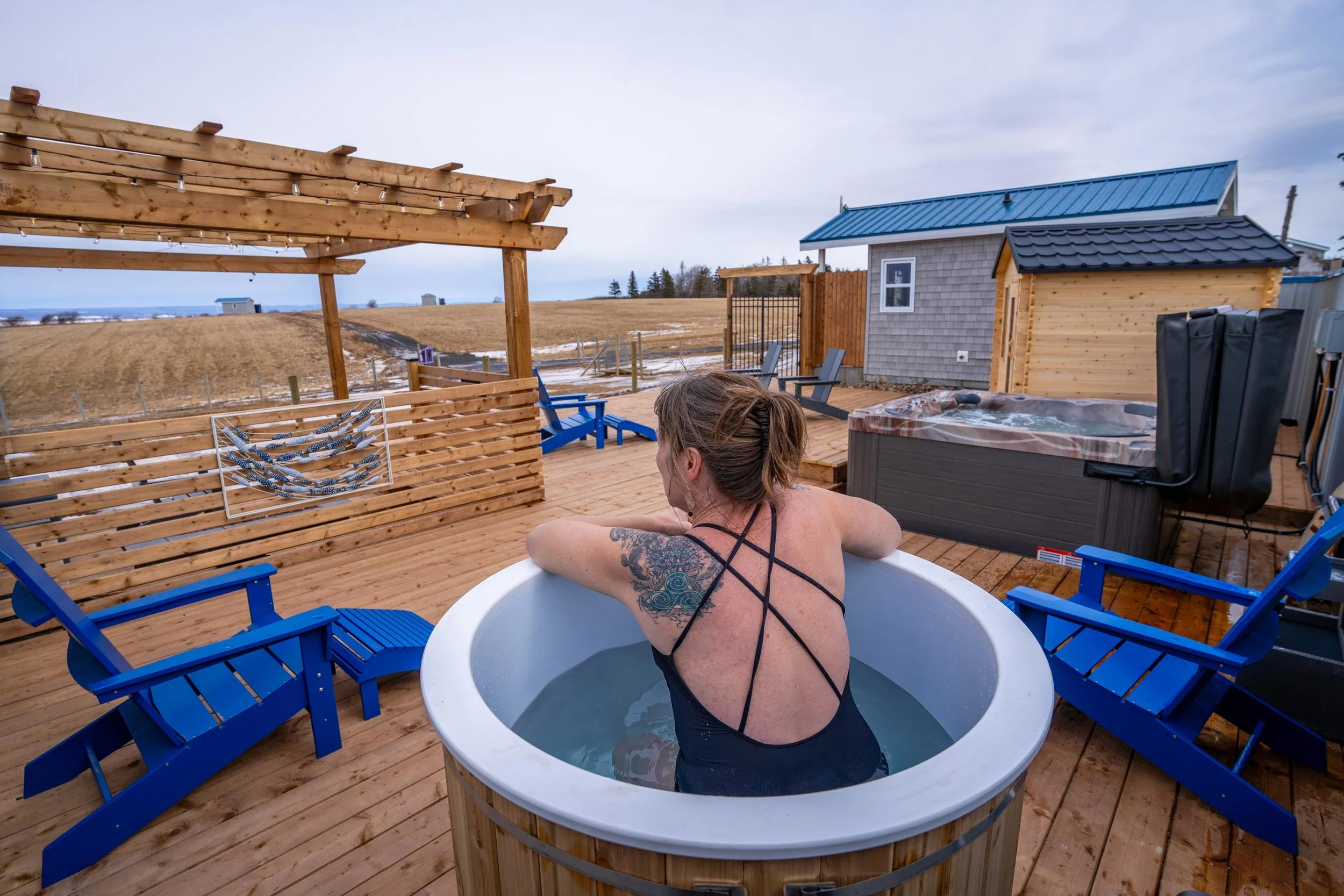 Person relaxing in a hot tub on a wooden deck outdoors, with a landscape of fields and other cabins at Ravens Rest Retreat