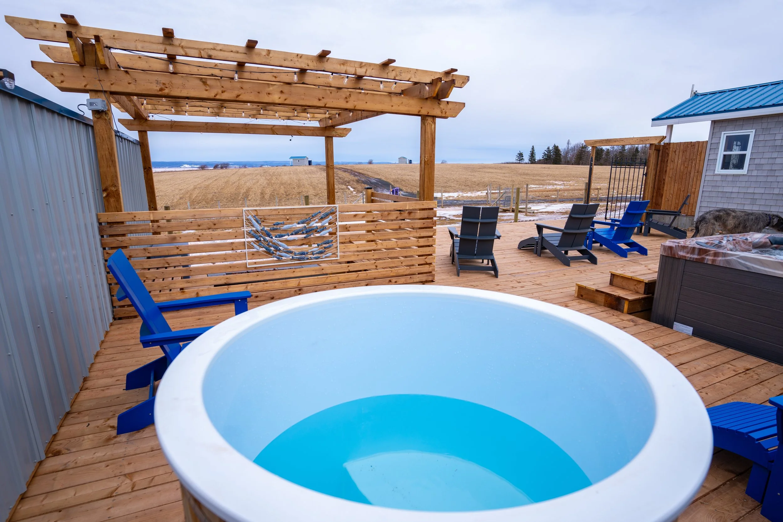 Wooden deck with blue chairs, cold plunge tub, and a patio cover overlooking open farmland.