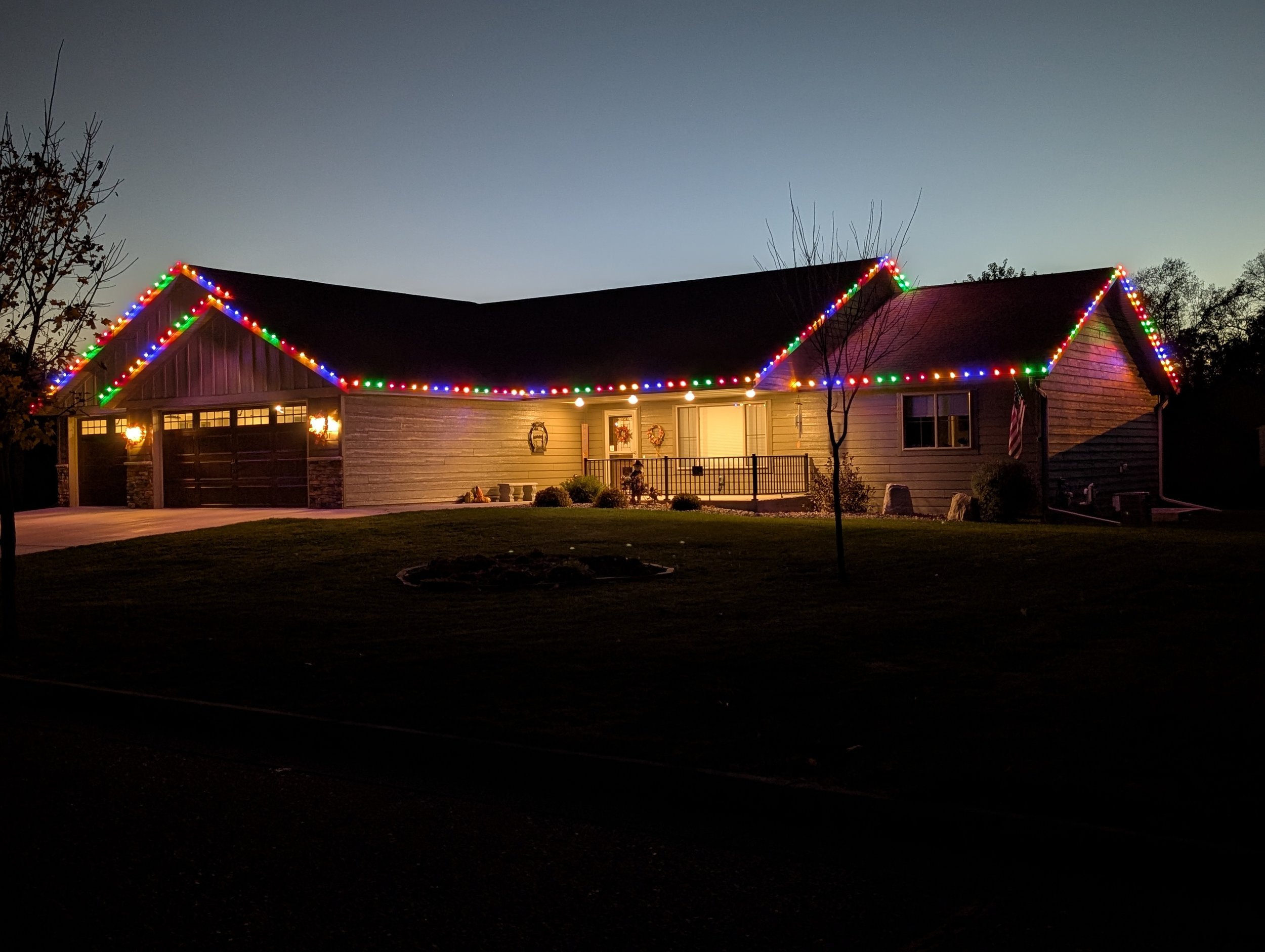 A house decorated with colorful string lights along the roofline during dusk or early evening, with lit porch lights, trees, and a well-maintained front yard.