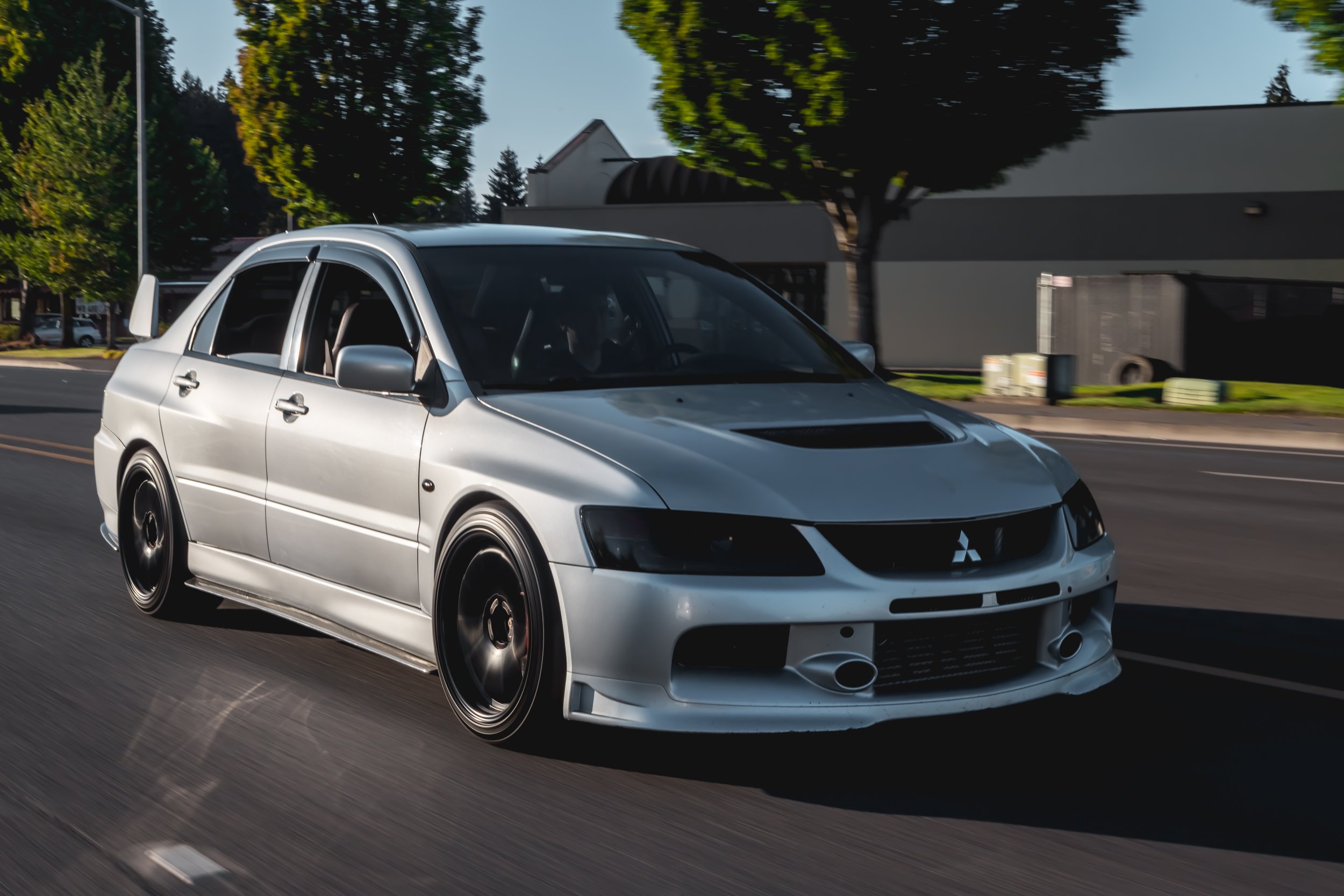 A silver Mitsubishi Lancer evolution car racing on a city street, with trees and buildings in the background.