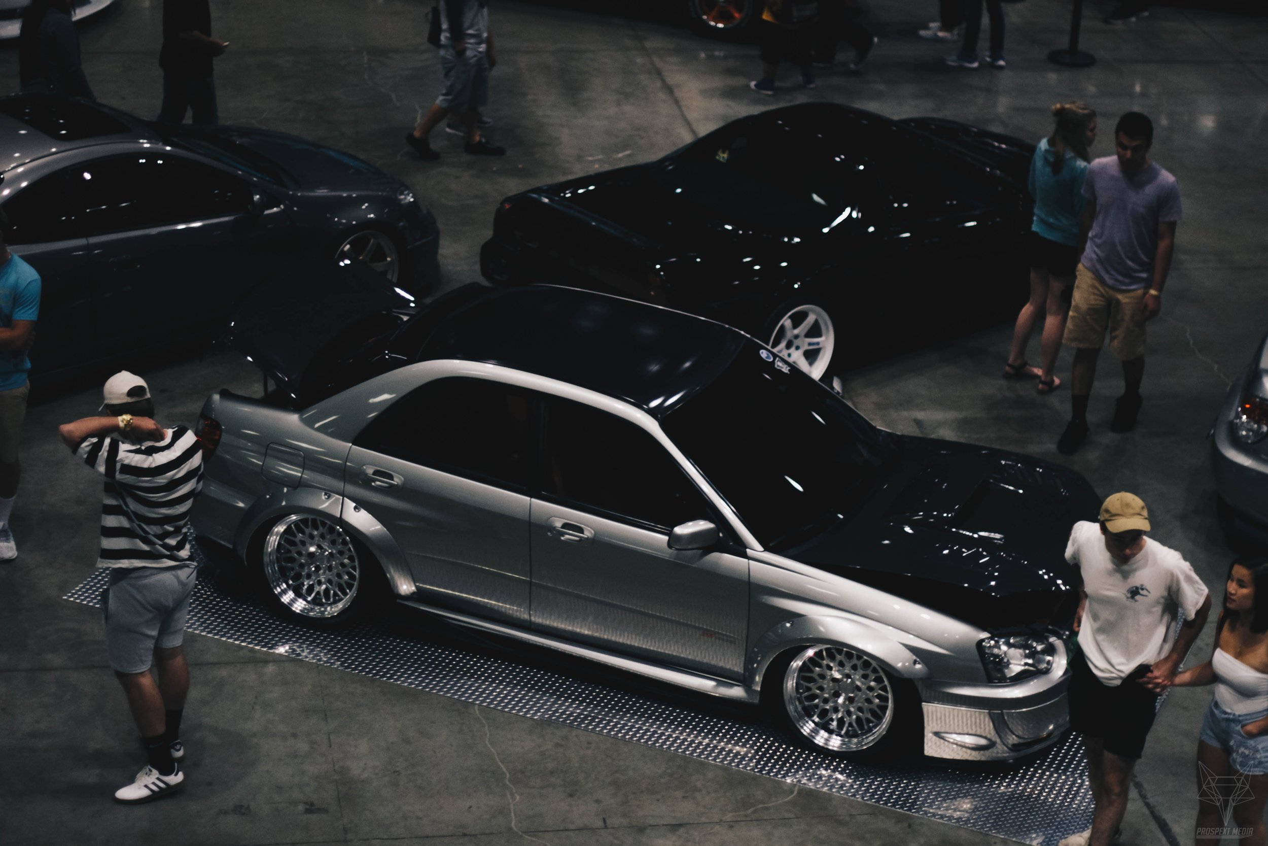 A silver and black modified car with custom rims is on display at an indoor car show, surrounded by people.