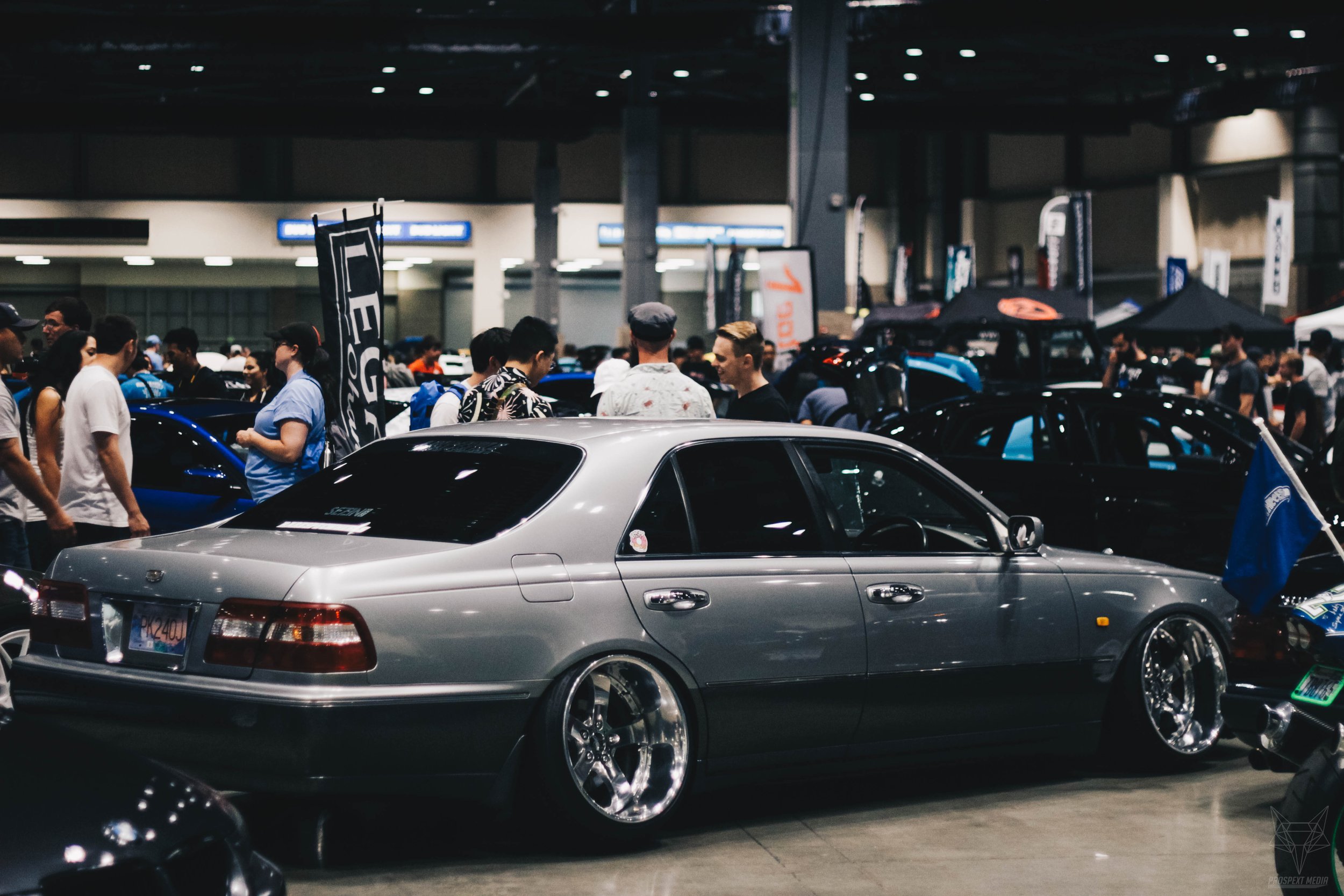 A silver lowered luxury sedan with aftermarket rims on display at a car show, surrounded by people and other cars under bright indoor lights.