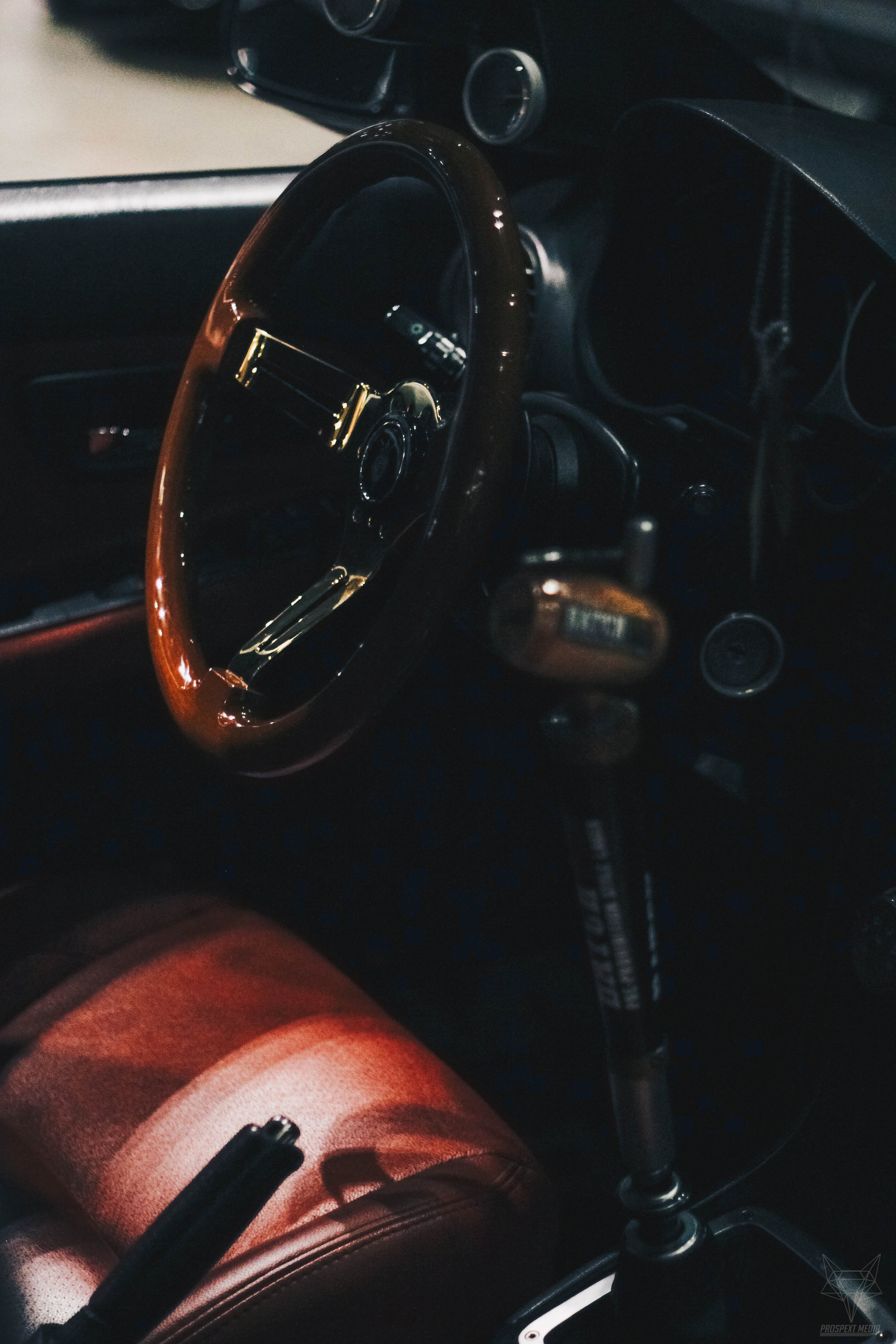 Interior of a car showing a wooden steering wheel, a gear shift, and a brown leather seat.