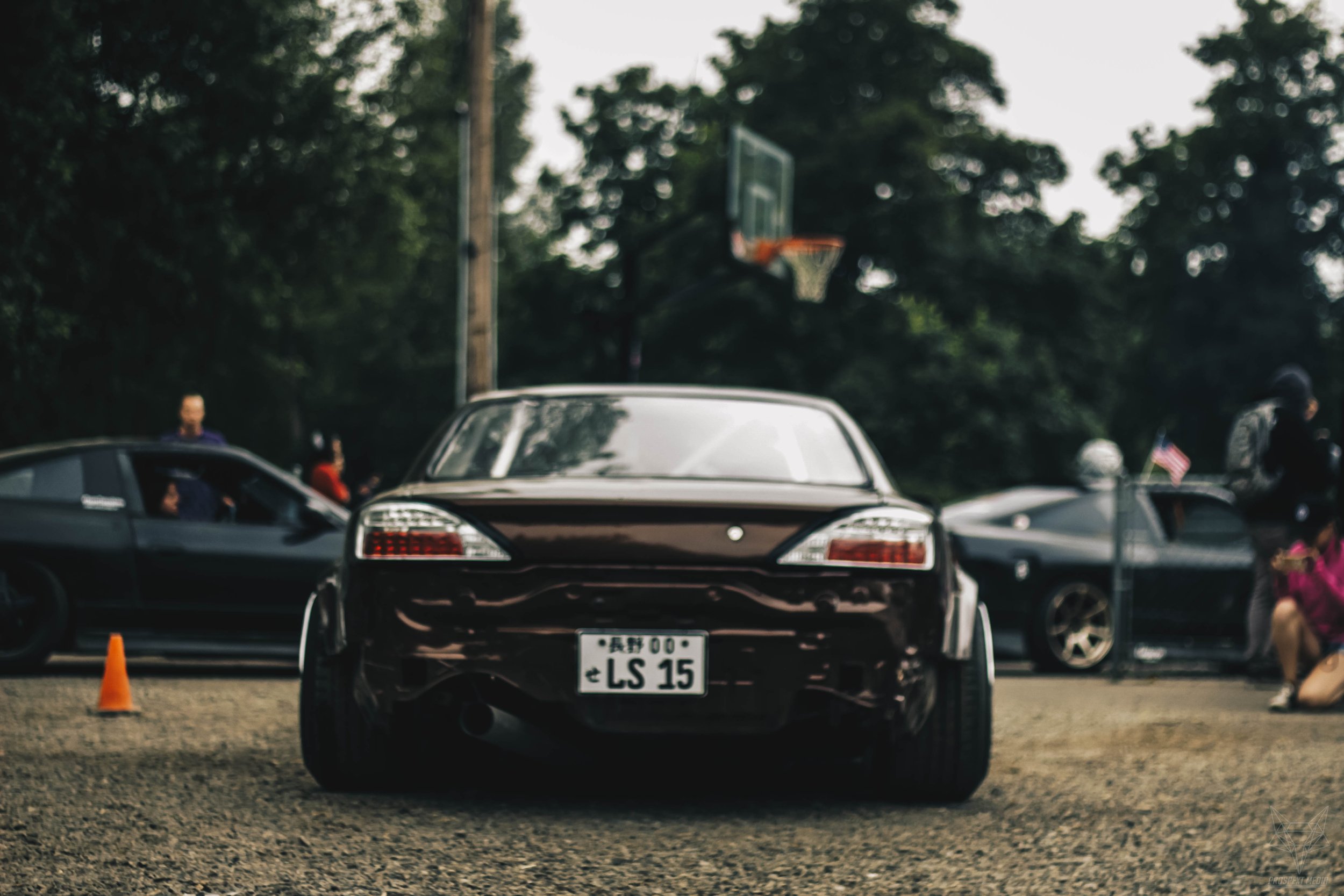 Rear view of a black sports car with Japanese license plate, parked on gravel with other vehicles and people around, basketball hoop in the background, outdoor setting with trees.