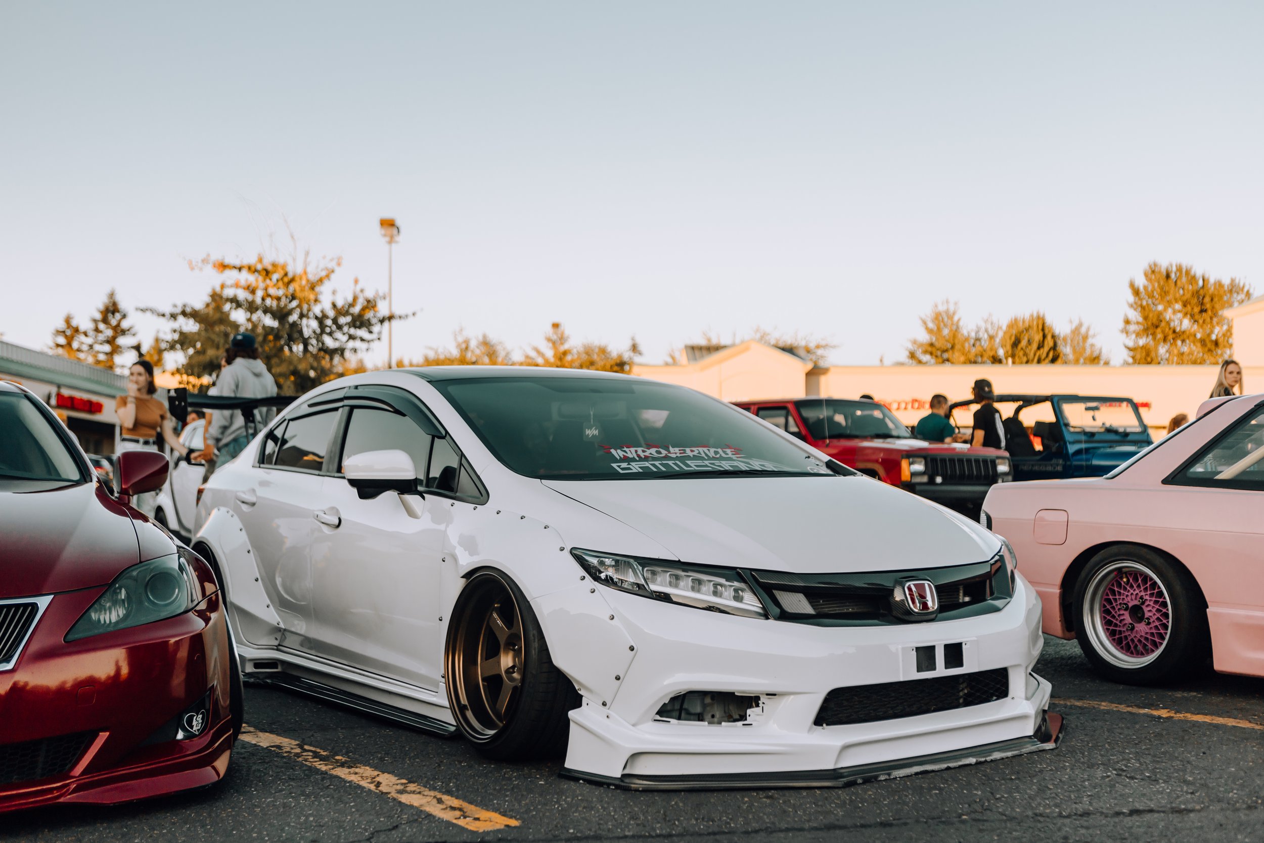 White modified Honda Civic with wide body kit, gold wheels, and lowered stance at a car show, with other cars and people in the background.