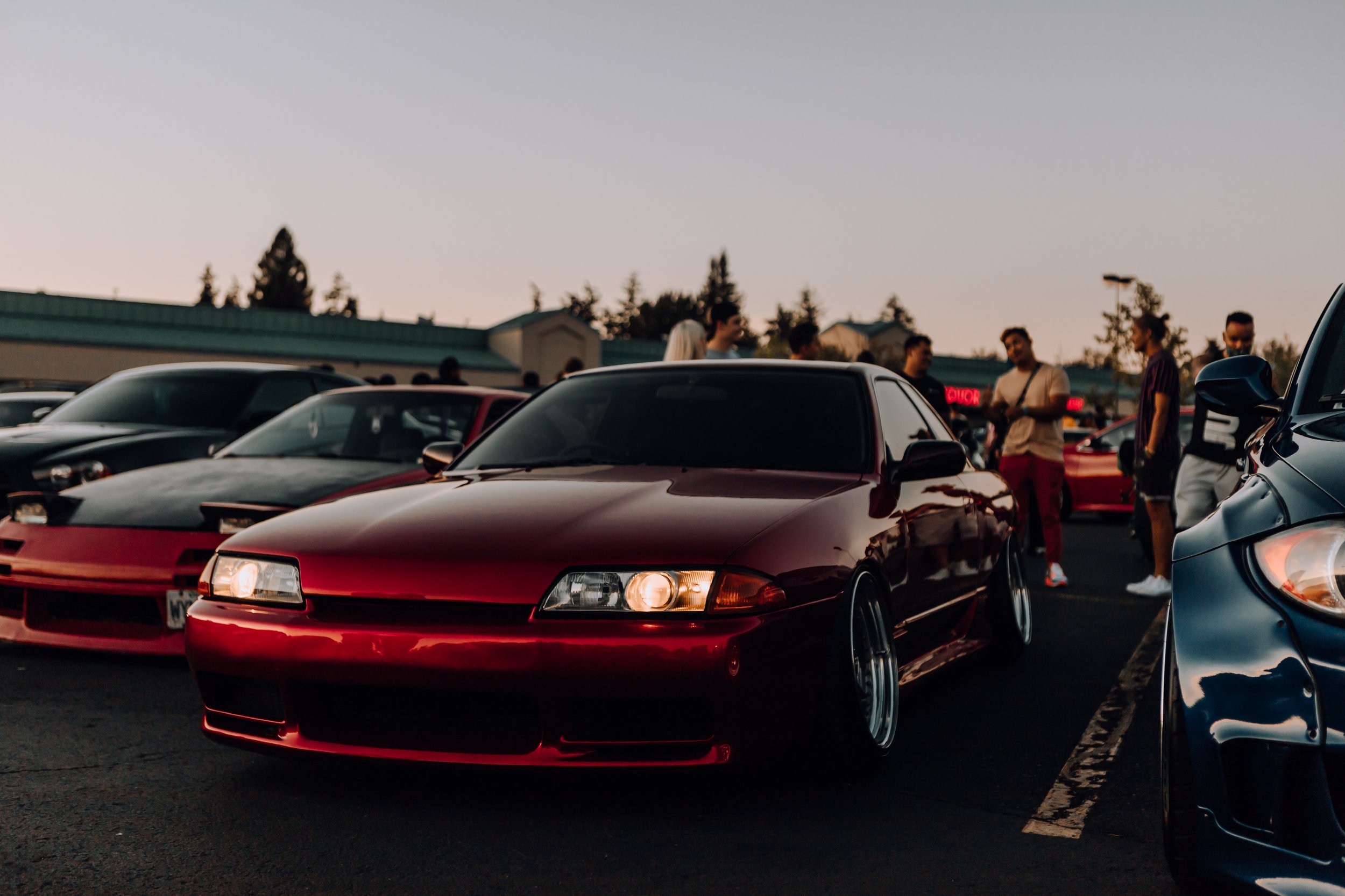 Red sports car parked at a car meet with people standing around in the background during sunset.