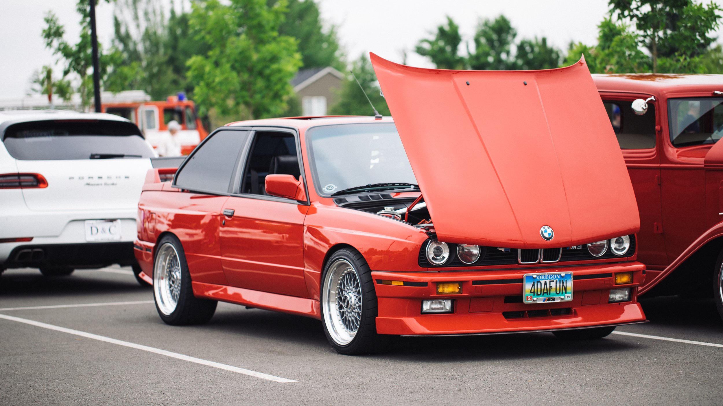 Red BMW with open hood, parked at a car show, surrounded by other vehicles including a white Porsche and a vintage red truck, with green trees in the background.