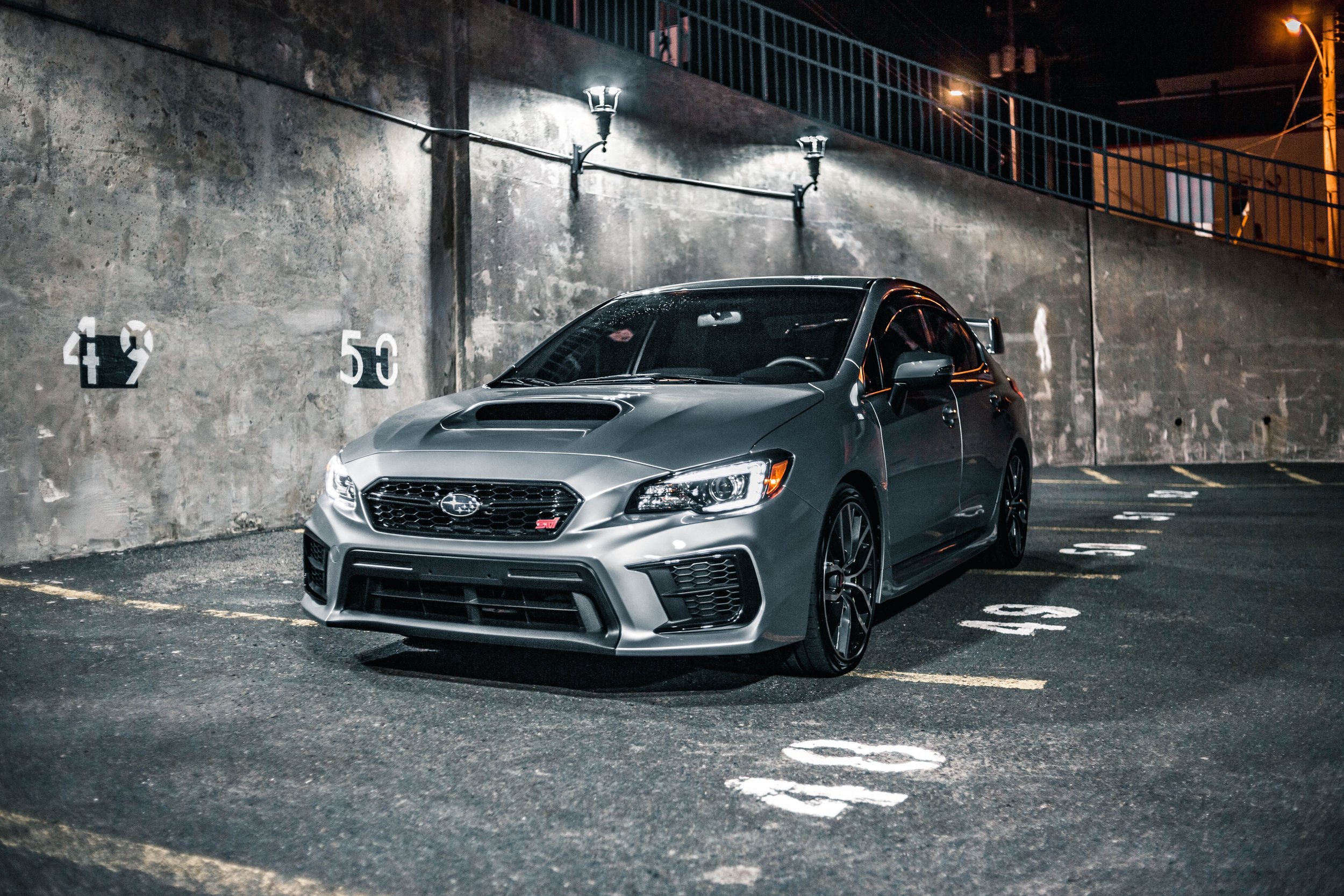 Gray Subaru WRX STI parked in an outdoor parking lot at night, with parking space numbers 48, 49, 50, and 51 visible on the ground. The setting is illuminated by streetlights, and a concrete wall with lighting fixtures is in the background.
